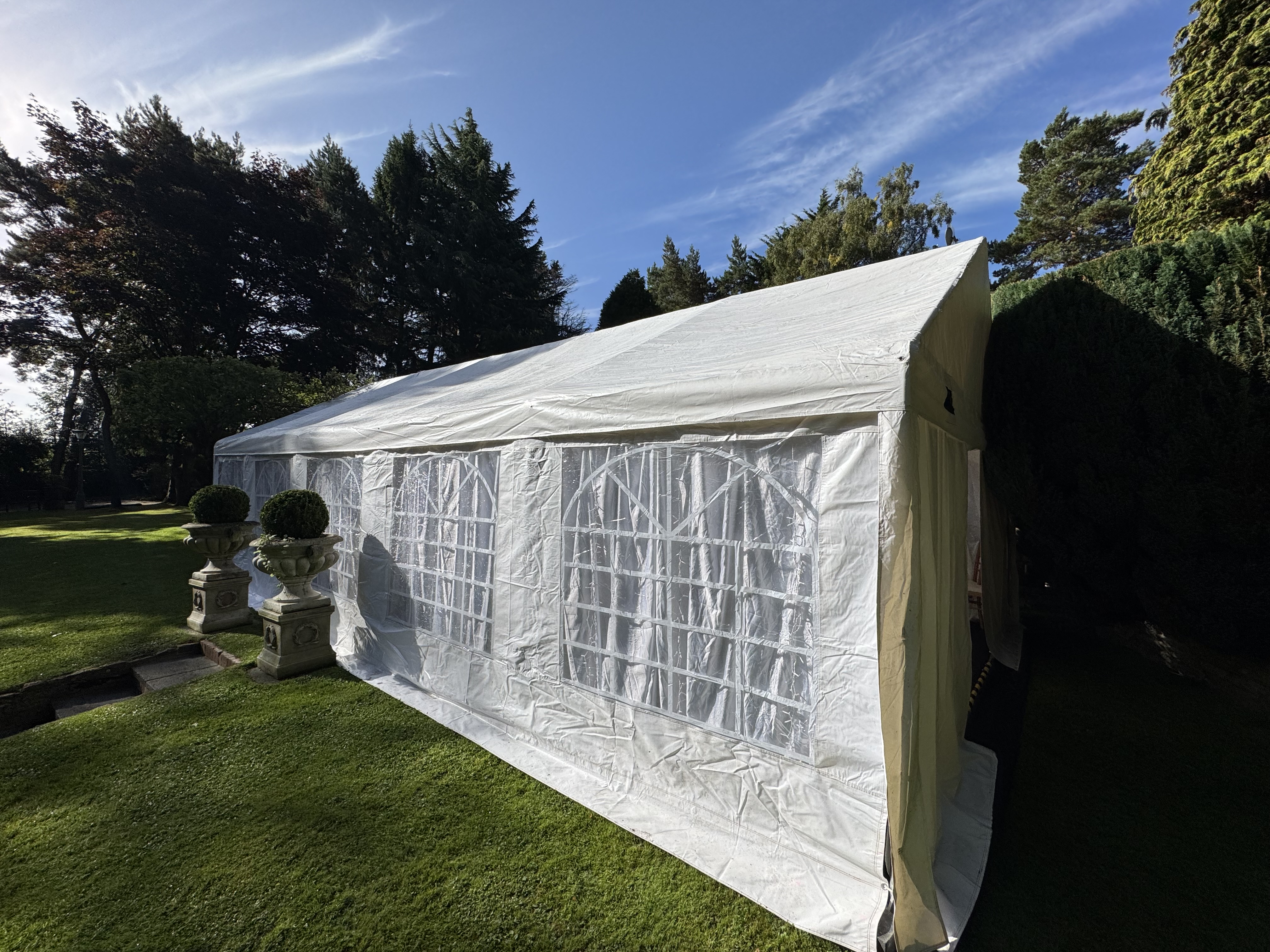 A spacious white tent with elegant window panels stands on lush green grass, surrounded by decorative topiary, under bright daylight.