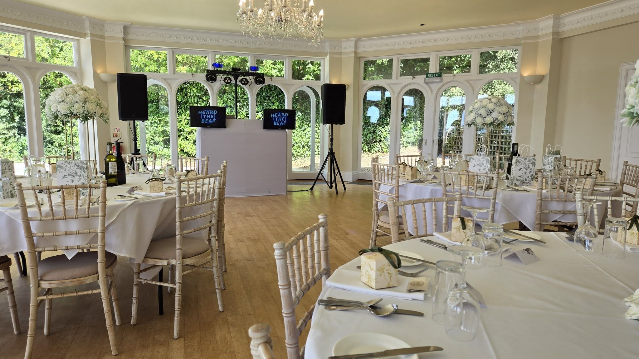 A bright, elegant venue features round tables draped in white linens, surrounded by polished chiavari chairs. Floral centerpieces enhance the charming atmosphere.