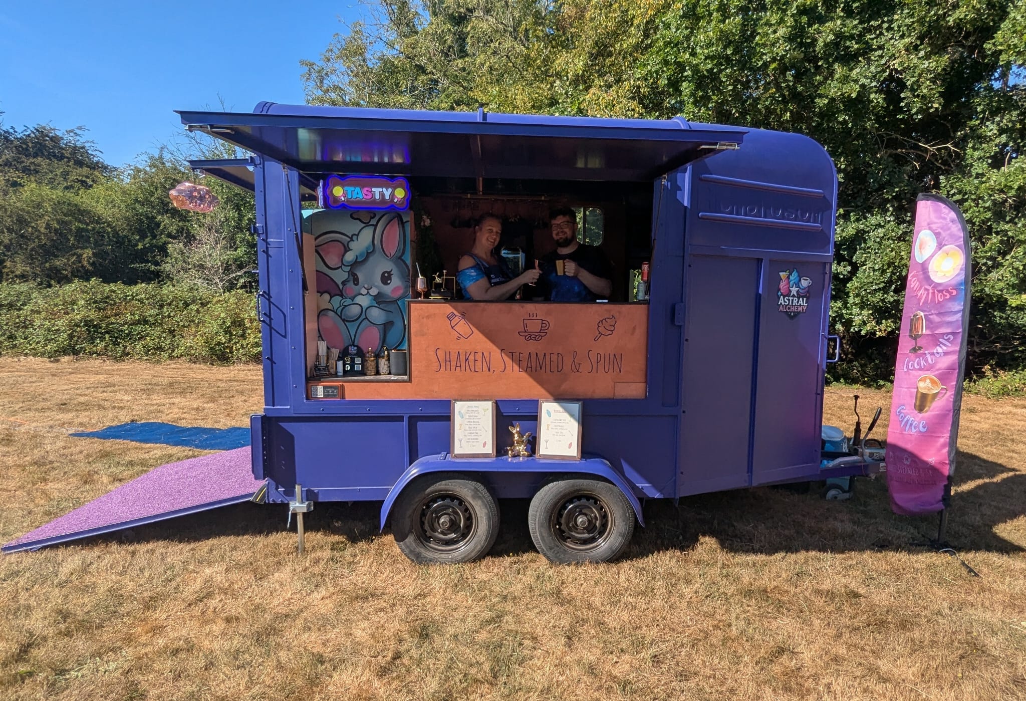 Purple cocktail catering van with open hatch, neon sign, and smiling staff serving drinks on a sunny lawn.