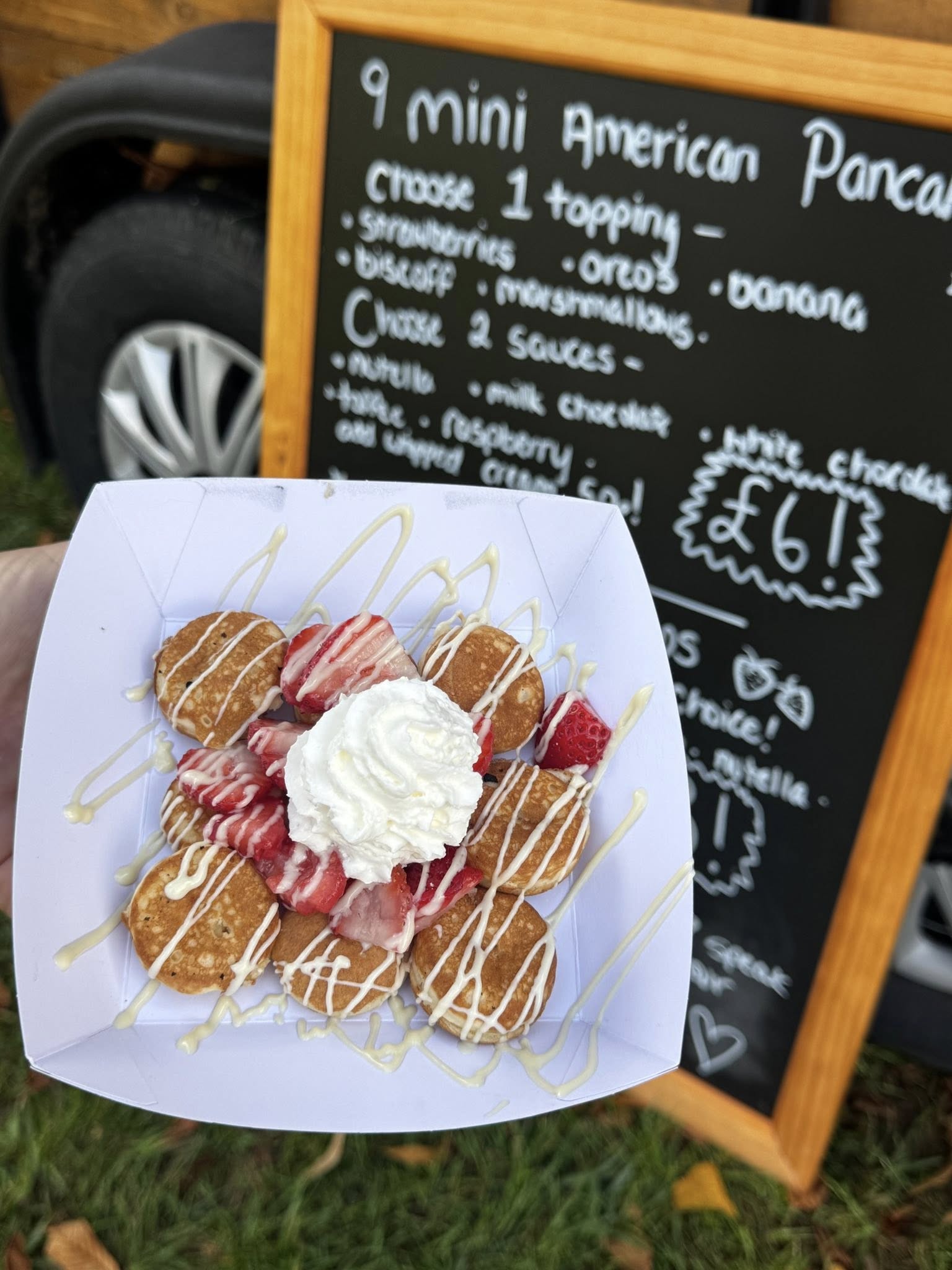 Nine mini American pancakes are beautifully arranged with strawberries and whipped cream, drizzled with white chocolate on a crisp white tray, set against a rustic wooden menu board in a vibrant outdoor atmosphere.