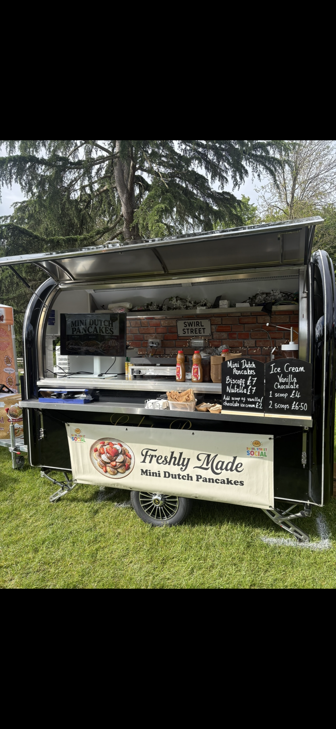 Open catering trailer with polished metal, brick backdrop, and chalkboard menus for mini Dutch pancakes and ice cream in bright daylight.