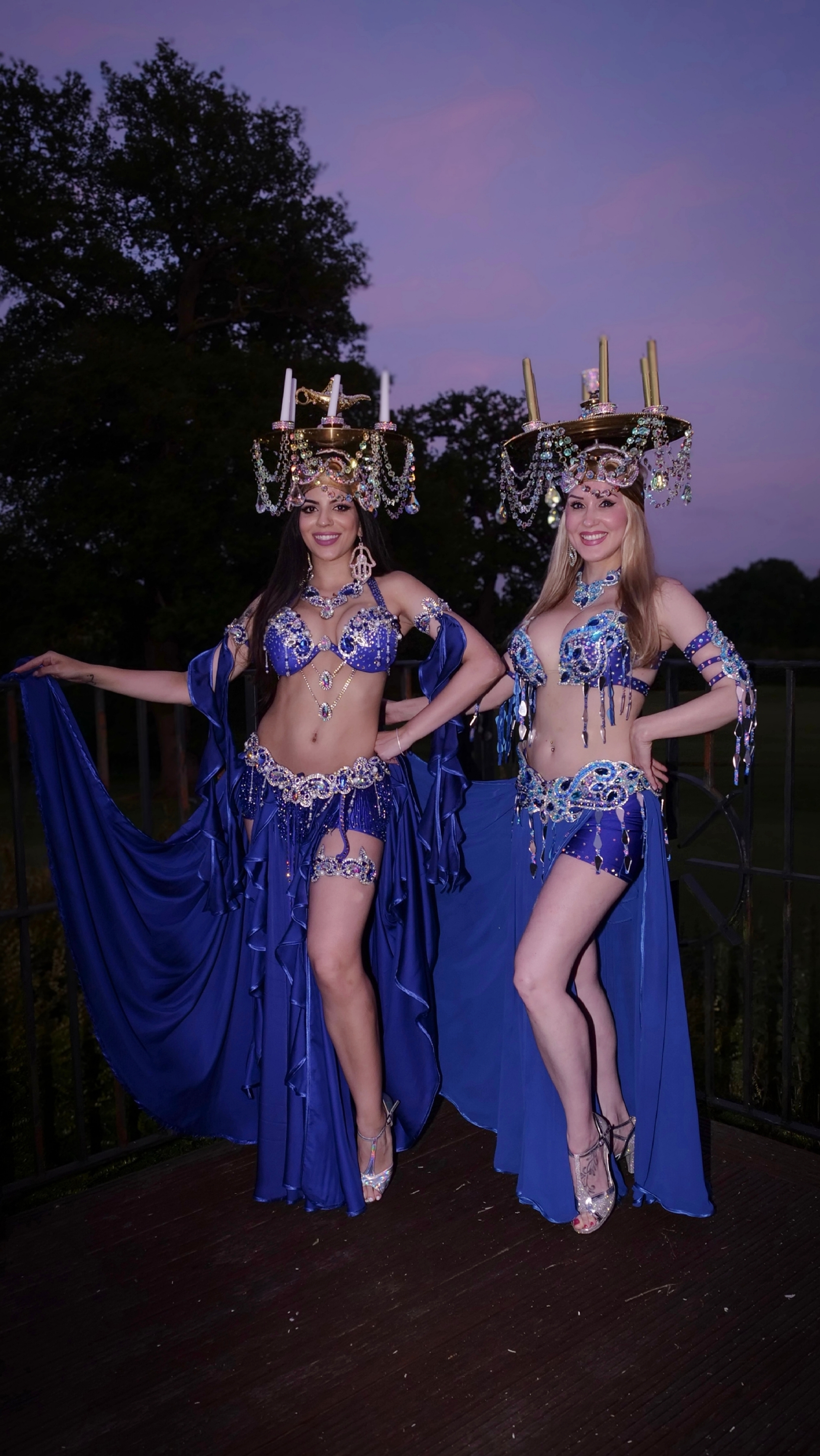 Two dancers pose in shimmering blue costumes adorned with jewels and flowing fabric, under a dusky sky, exuding vibrant energy.