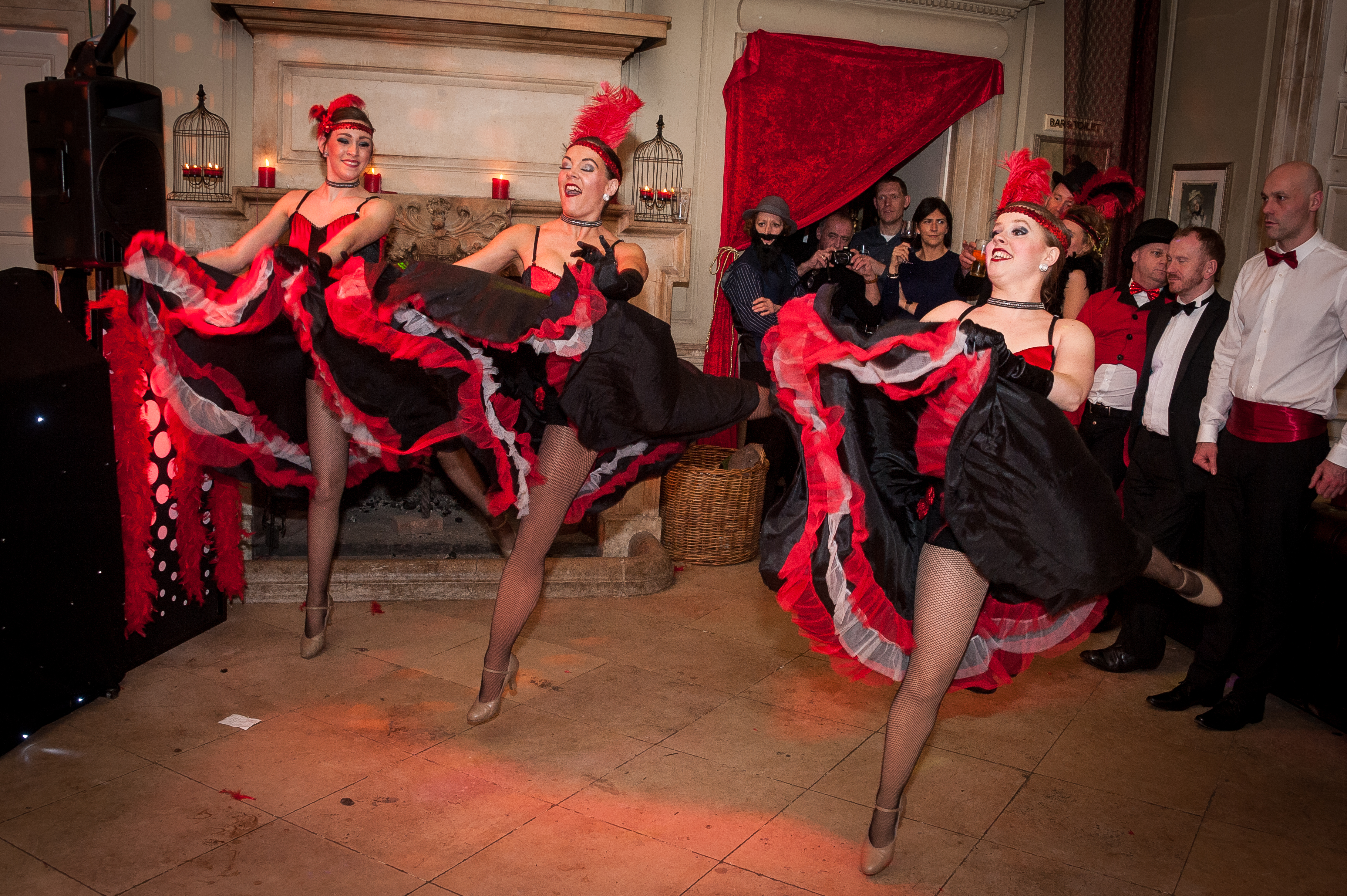 Cabaret dancers in red-and-black feathered costumes perform before a stone fireplace, with guests watching under warm candlelight at a chic event.