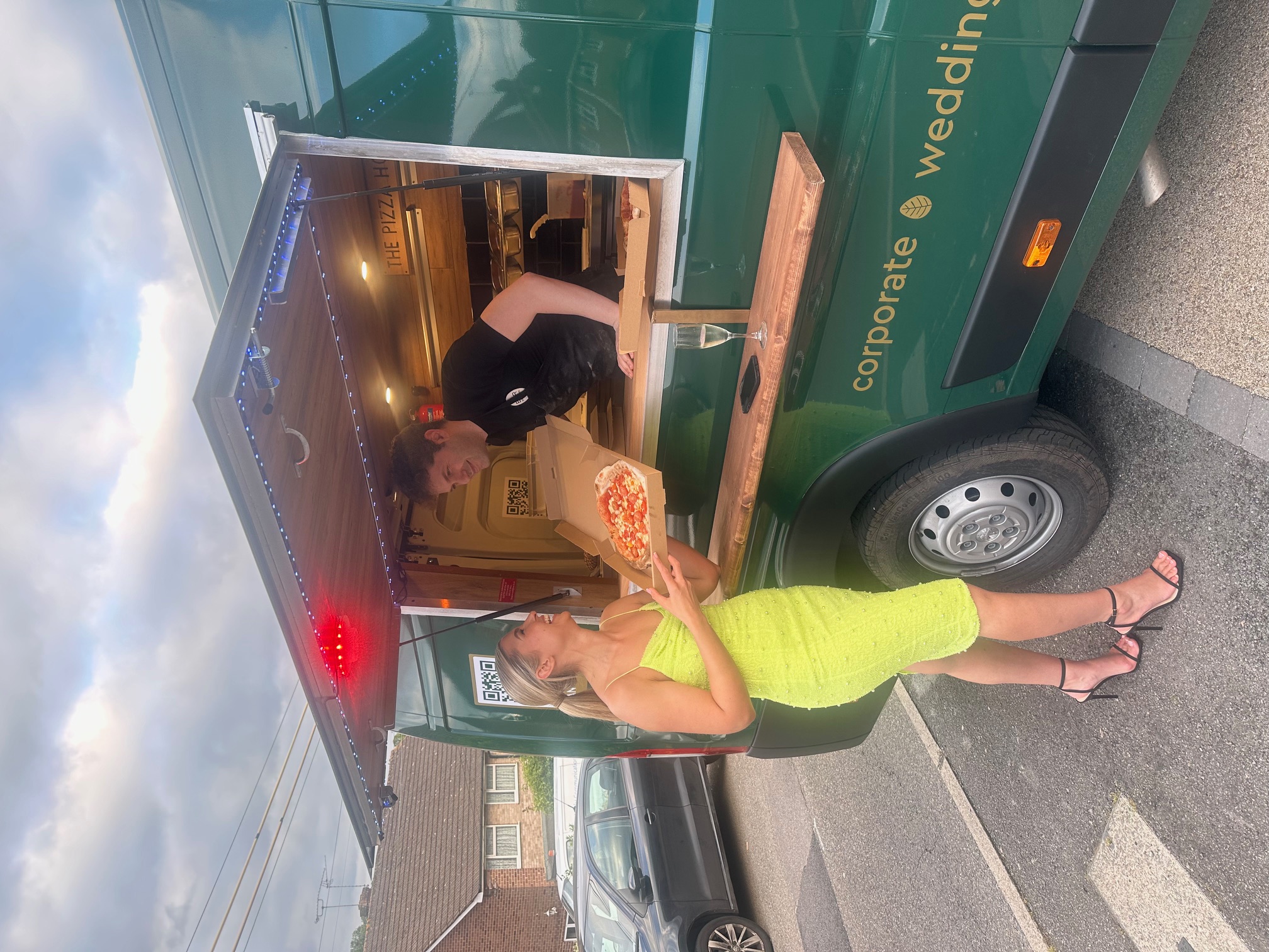 Bright catering scene at a green pizza van, with a neon-dress guest receiving a boxed pizza under warm LED lighting.
