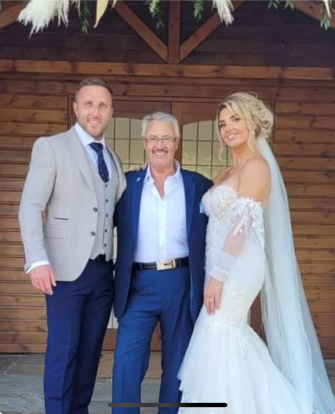 A bride in a lace gown and veil poses with two men outside a rustic wooden venue, radiating joyful wedding vibes under bright daylight.