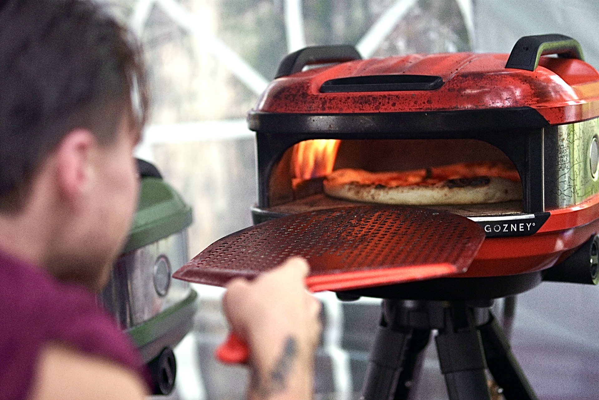 A vibrant red Gozney pizza oven, emitting warmth, showcases a golden-brown pizza being pulled out, set against a rustic backdrop, highlighting outdoor catering.