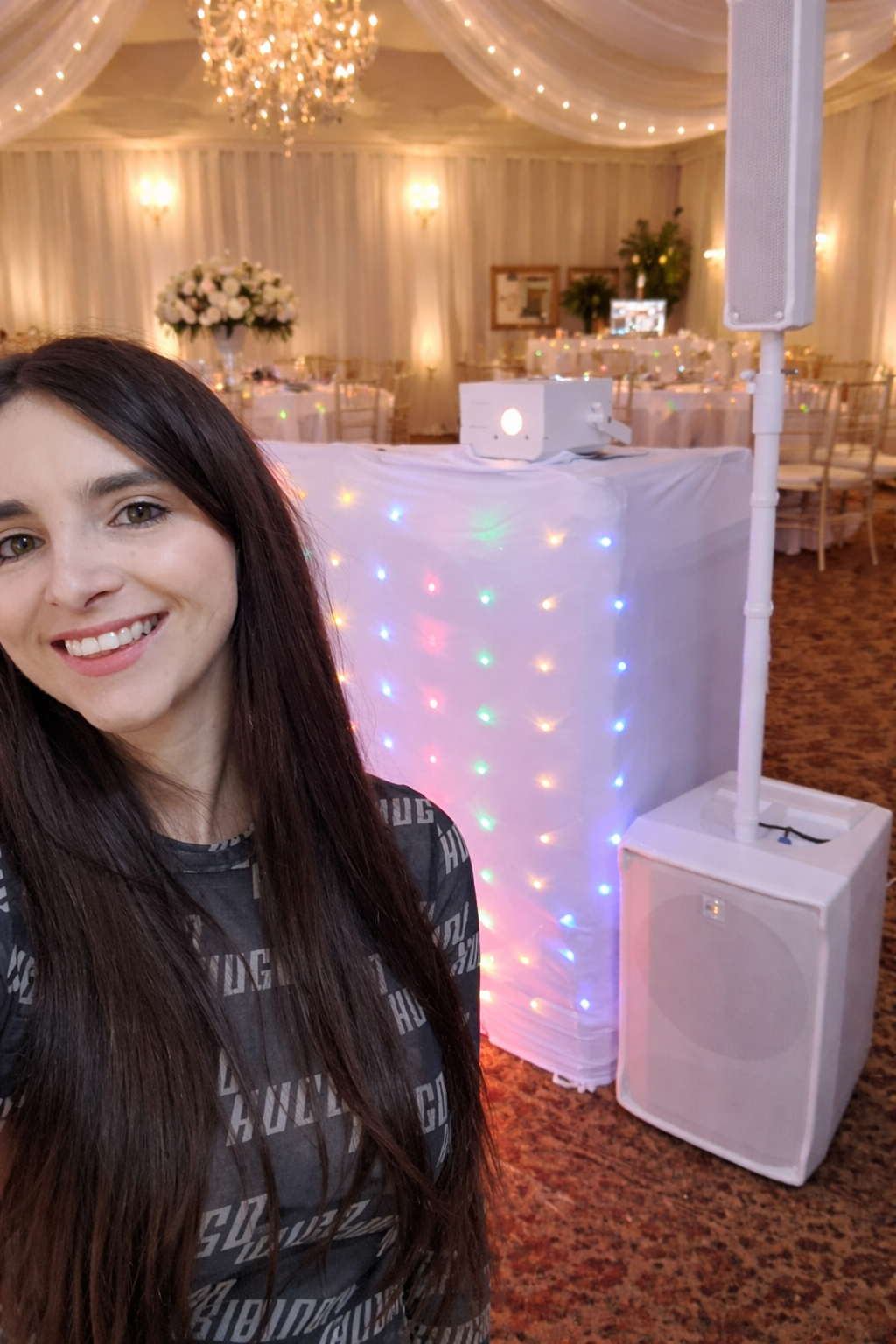 A beautifully decorated banquet hall features elegant white drapes and soft lighting, with a vibrant audio setup beside a cheerful host.