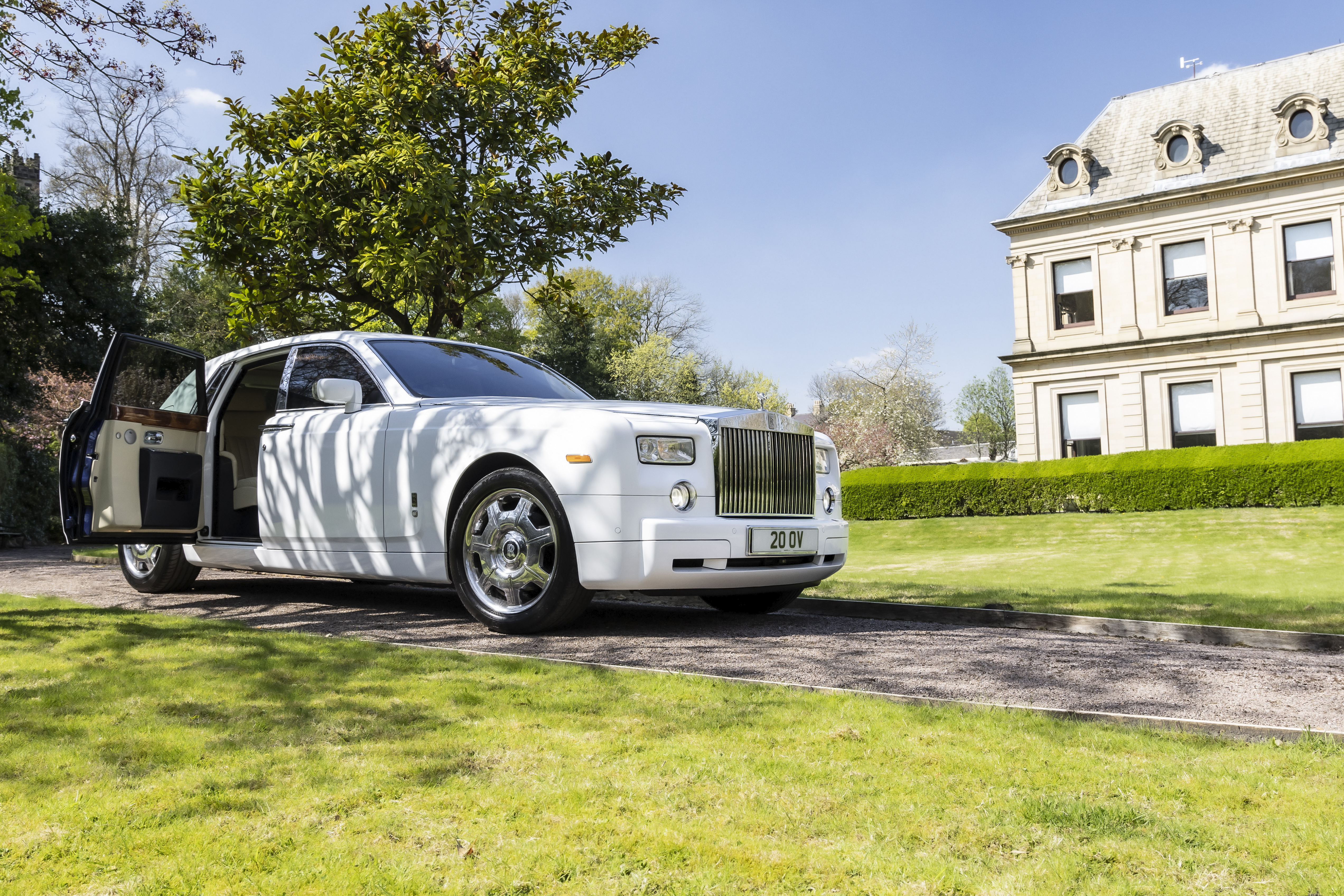 A pristine white Rolls-Royce is parked elegantly on a gravel path, framed by lush green grass and a grand building, exuding luxury.