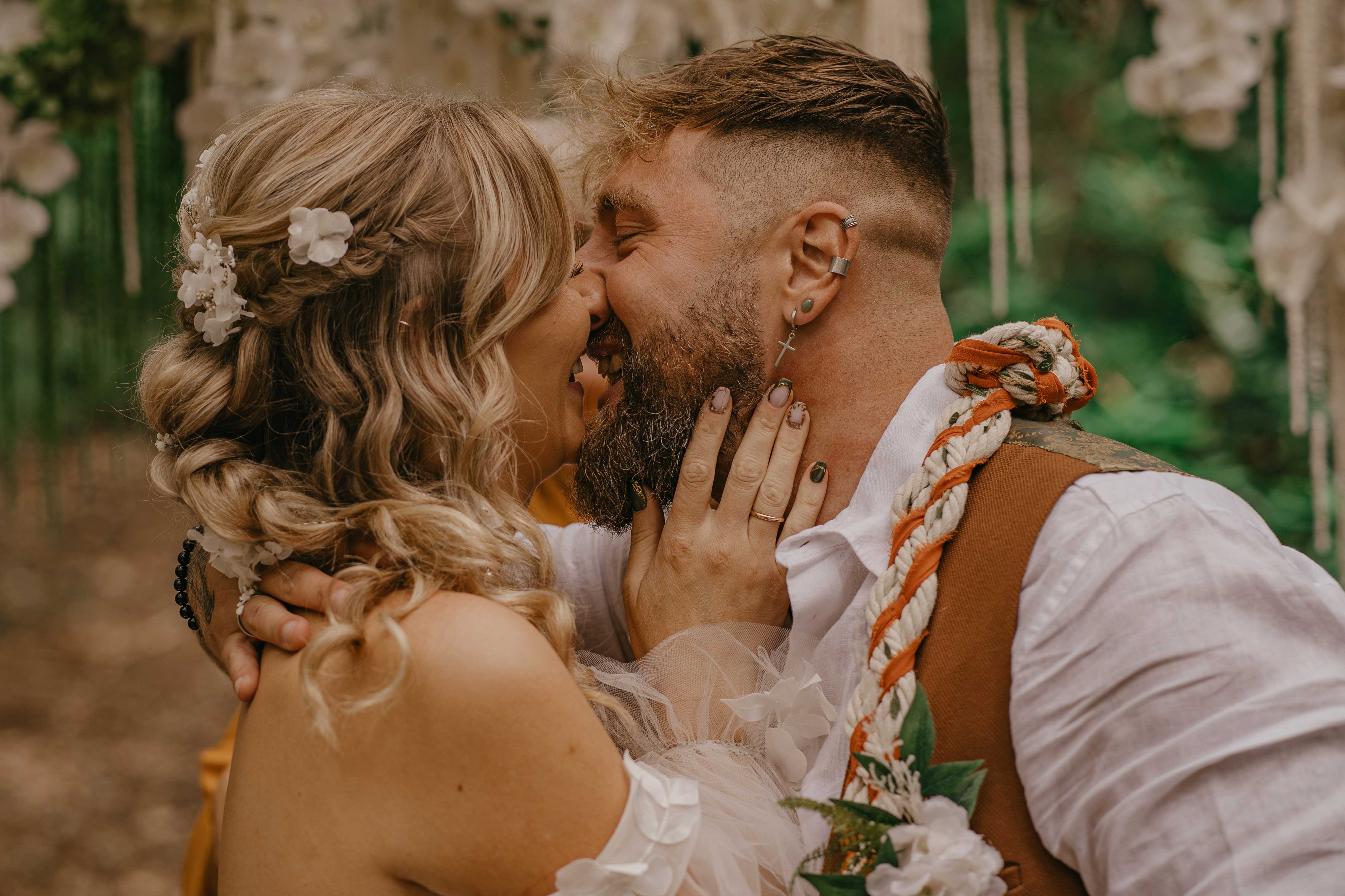 Intimate boho wedding close-up: couple kissing amid white floral arch, warm soft daylight, bridal hair flowers, polished styling, romantic vibe.