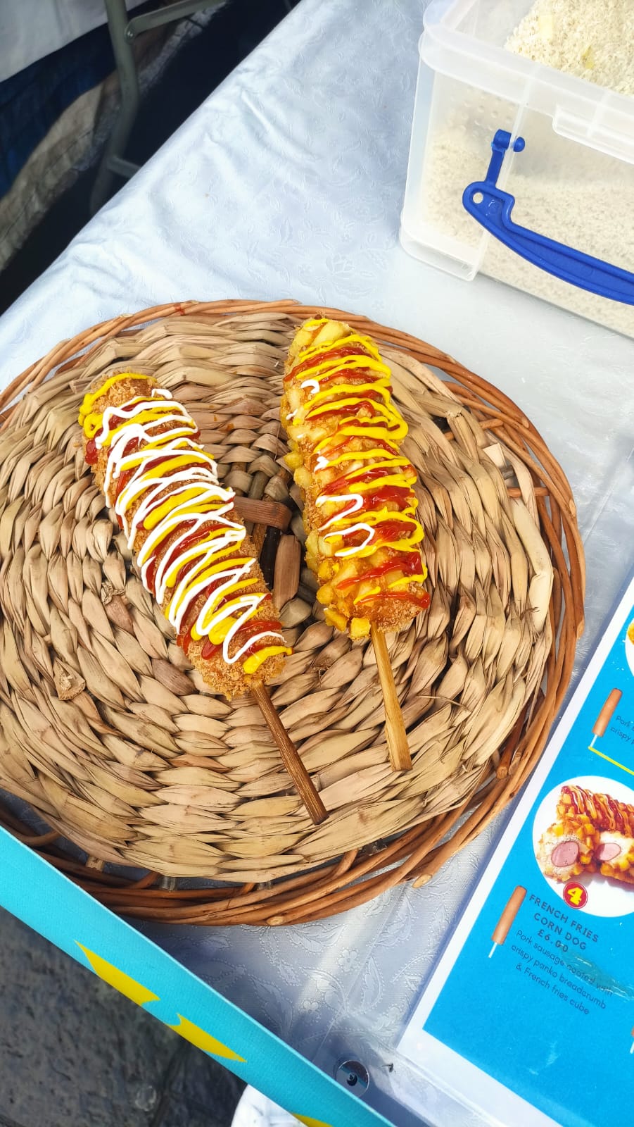 Two loaded corn dog skewers with ketchup, mustard and mayo rest on a woven wicker tray, bright catering display.