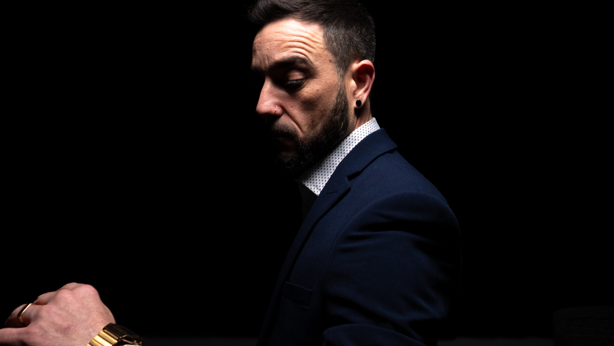Moody studio portrait of a bearded man in a navy suit, dramatic side lighting, black backdrop, crisp shirt detail, and polished elegance.