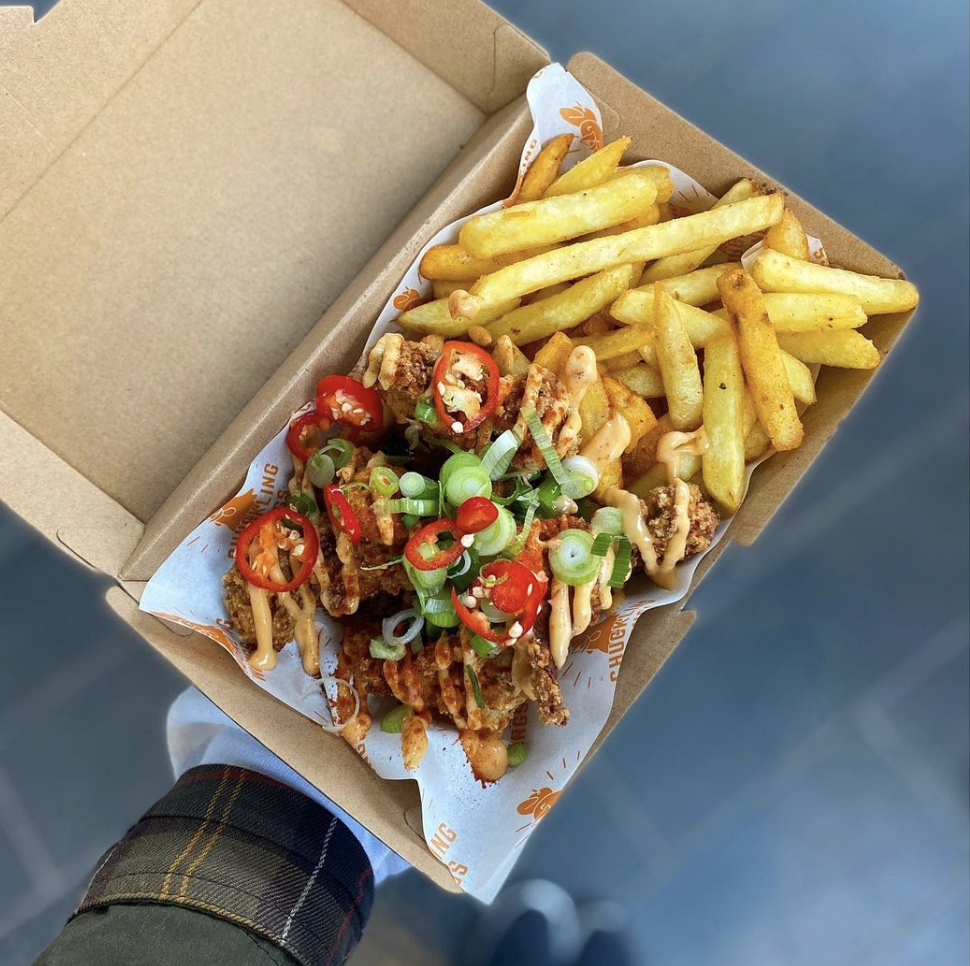 A cardboard box cradles crispy chicken topped with vibrant red chilies and scallions, alongside golden fries, showcasing delicious catering.