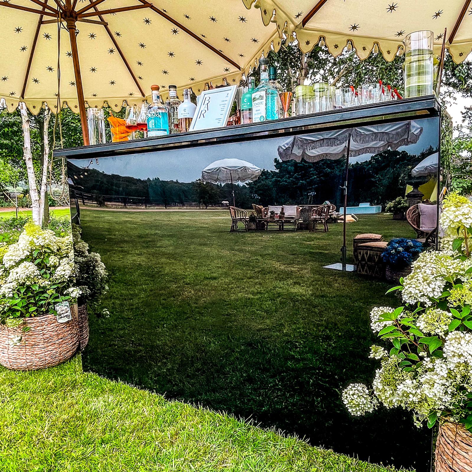 A sleek black bar displays a vibrant assortment of drinks, surrounded by lush greenery and elegant seating under a star-patterned umbrella, creating a festive ambiance.