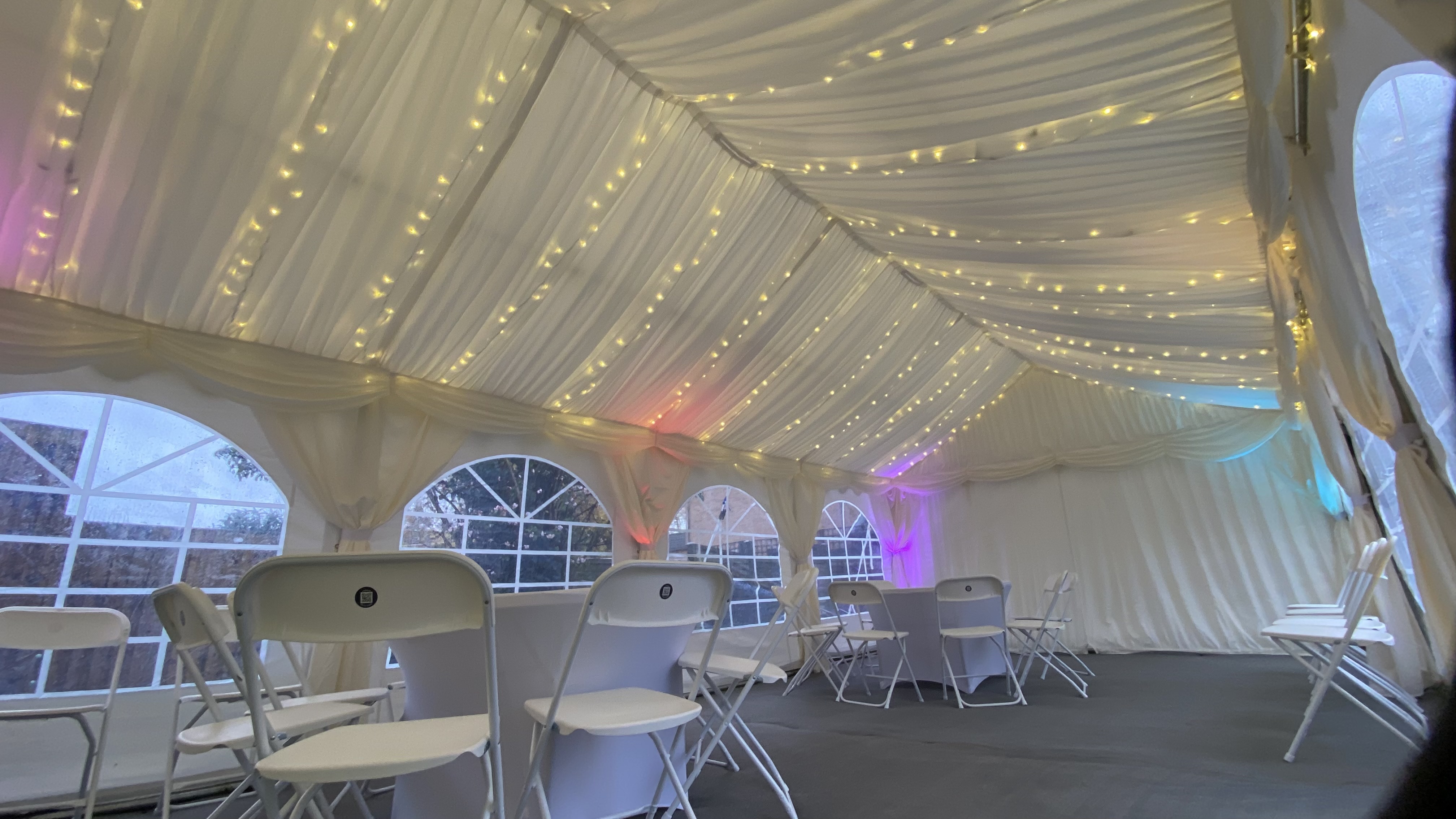 Elegant white draped wedding tent with fairy lights, arched windows, white folding chairs, cocktail tables, and soft colorful uplighting.