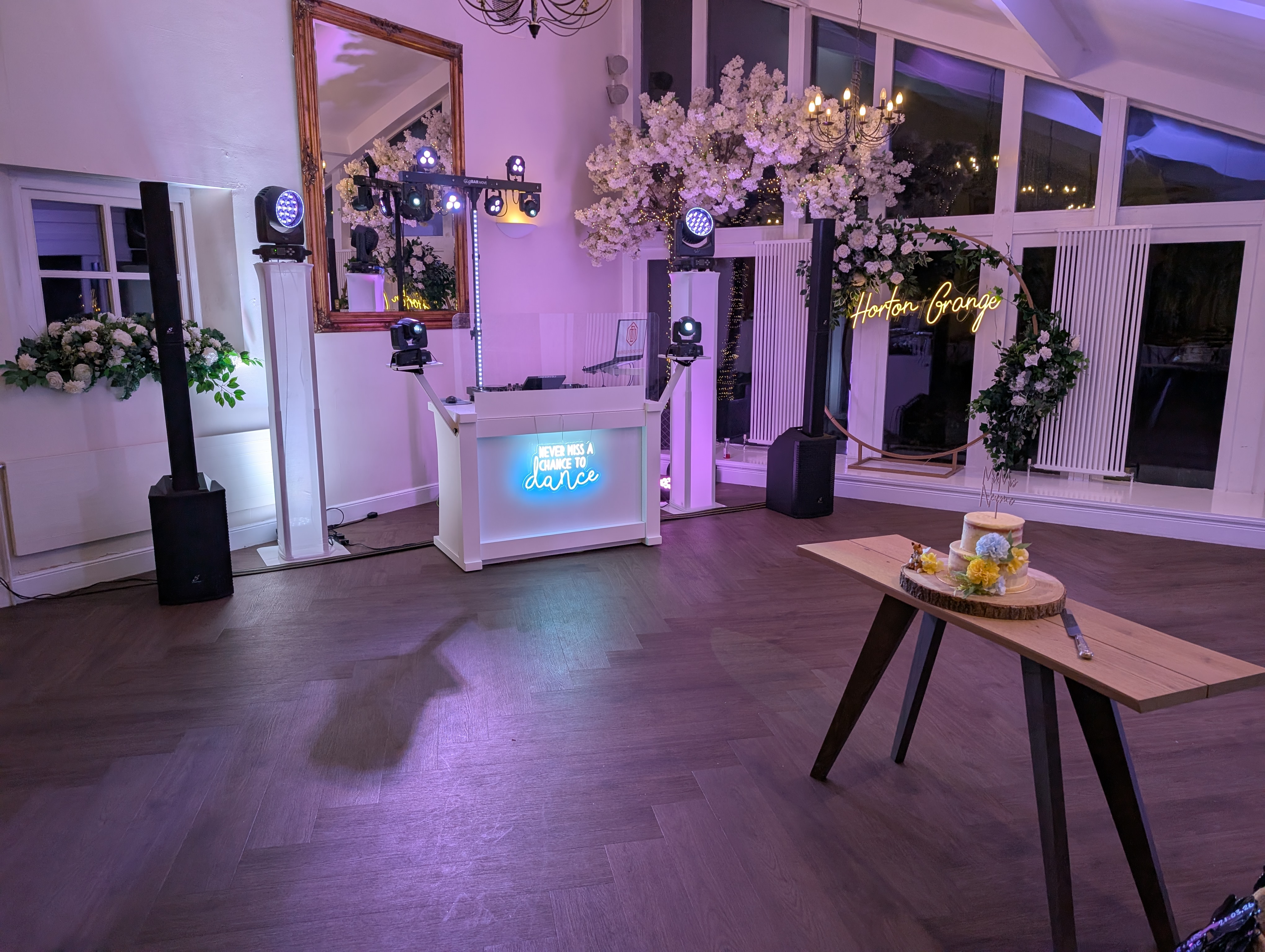 A chic event space featuring a vibrant DJ booth and elegant decor, with a wooden table displaying a floral cake under soft, colorful lighting.