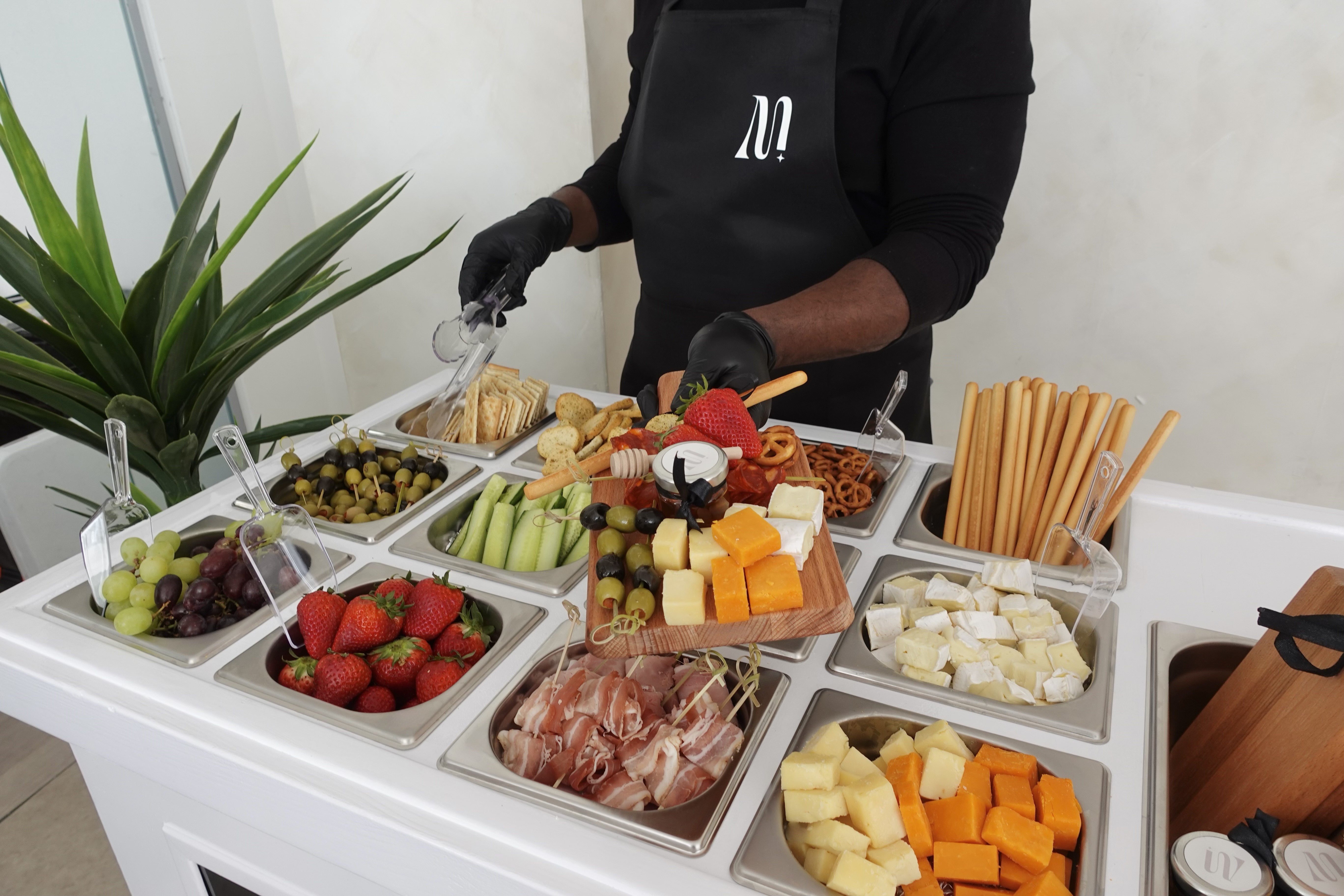 A vibrant buffet features fresh strawberries, assorted cheeses, olives, crackers, and pretzel sticks elegantly displayed in stainless containers under bright lighting.
