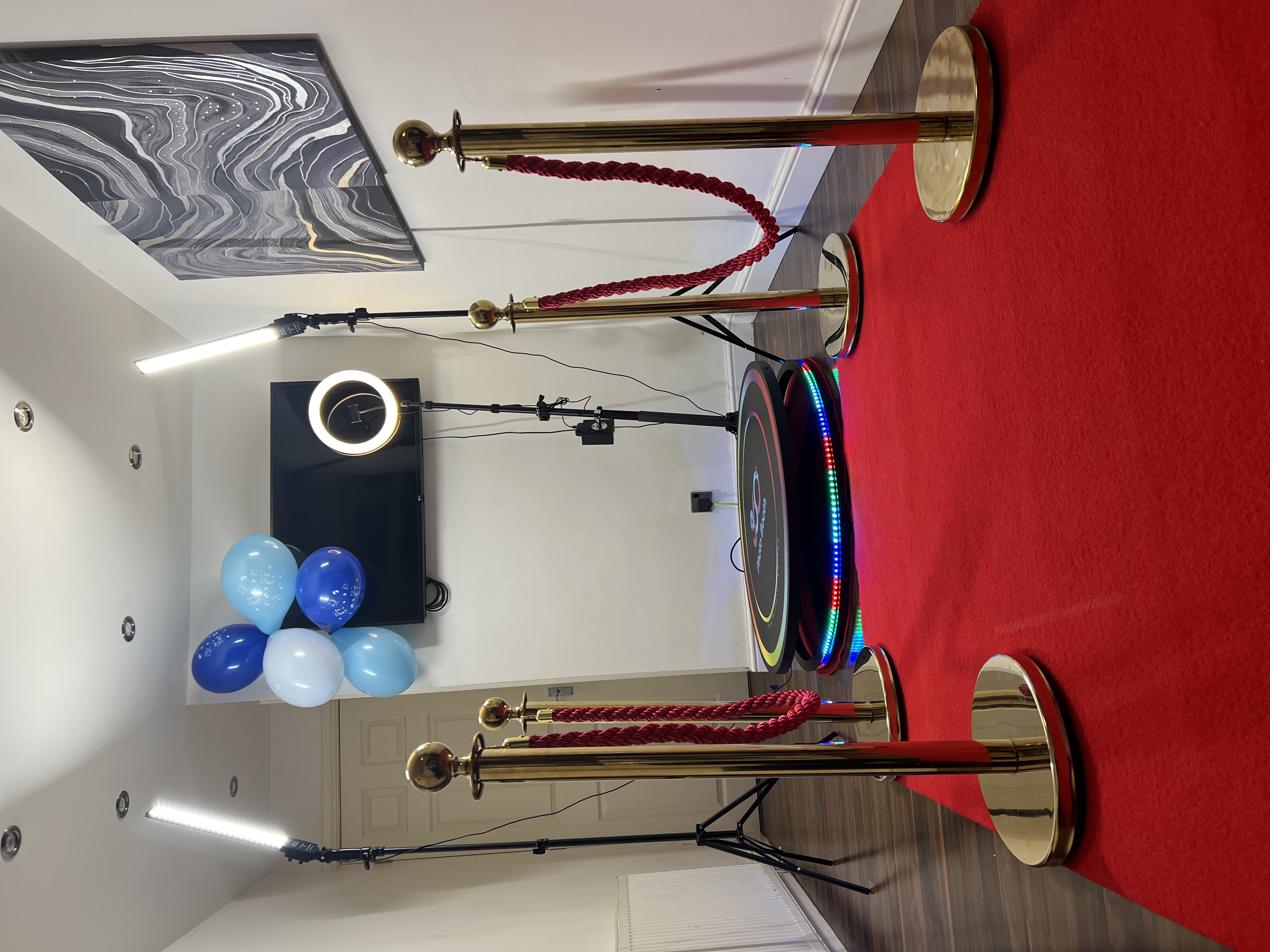 A vibrant red carpet leads to a circular illuminated platform, flanked by gold stanchions and balloons, creating a festive atmosphere.