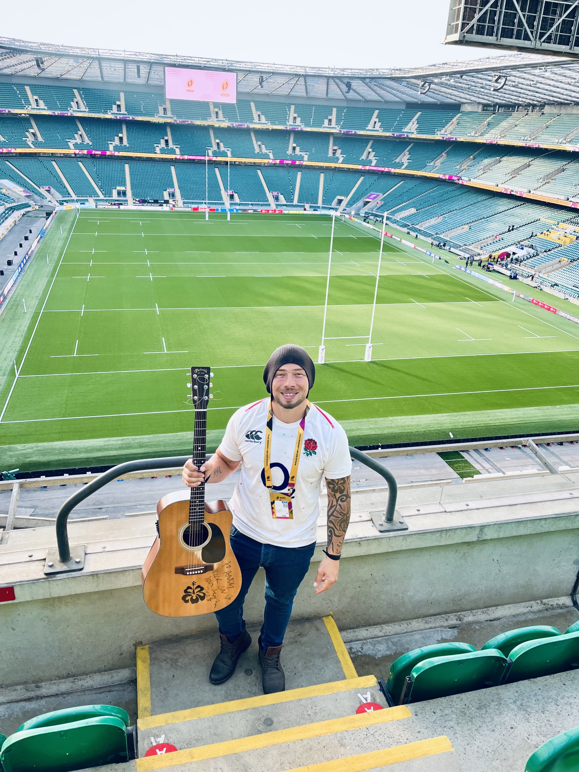 Smiling man with acoustic guitar on stadium steps, bright daylight, empty rugby field, teal seats, polished event venue atmosphere.
