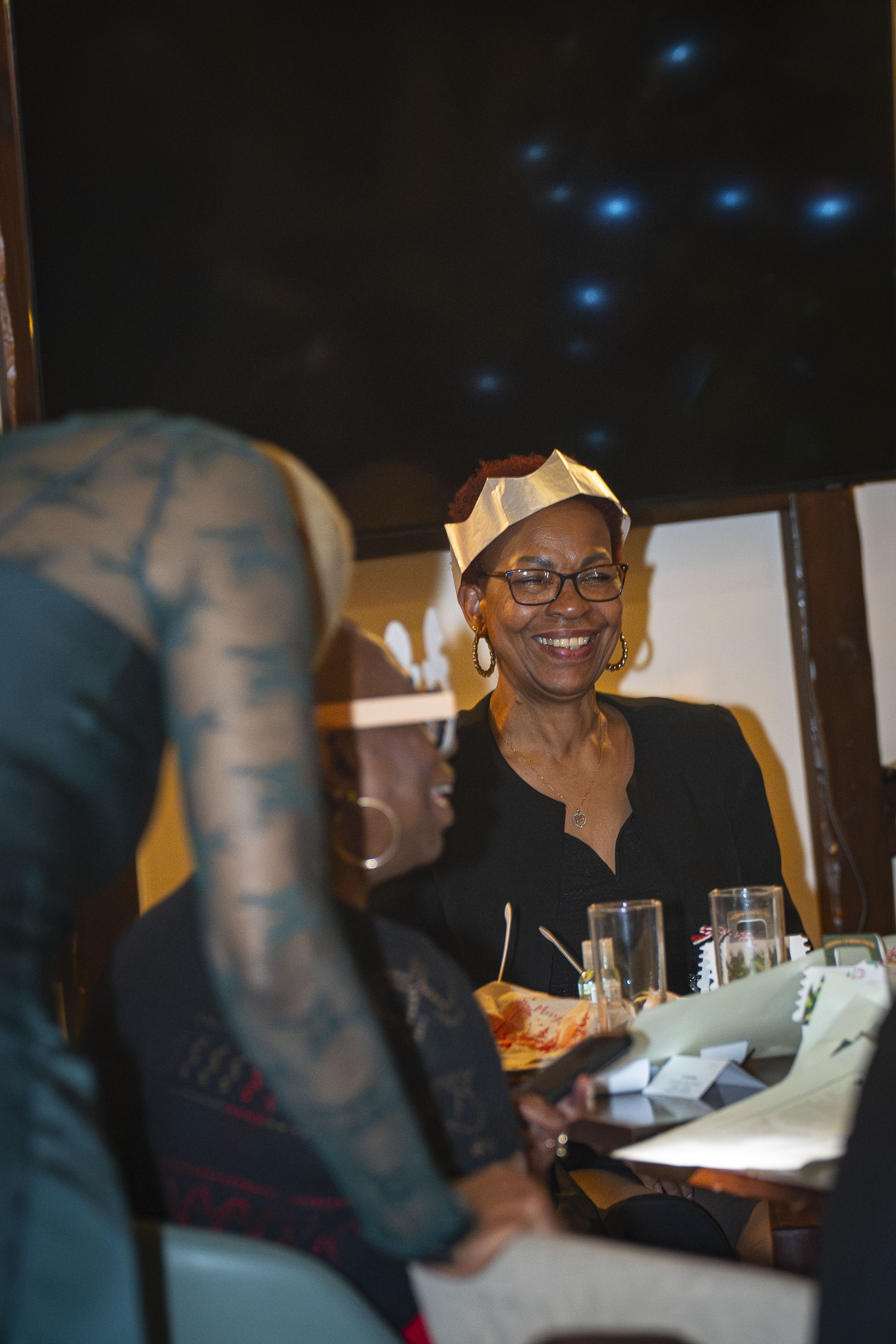A warmly lit gathering features cheerful guests, festive paper crowns, and drinks, embodying a lively celebration atmosphere.