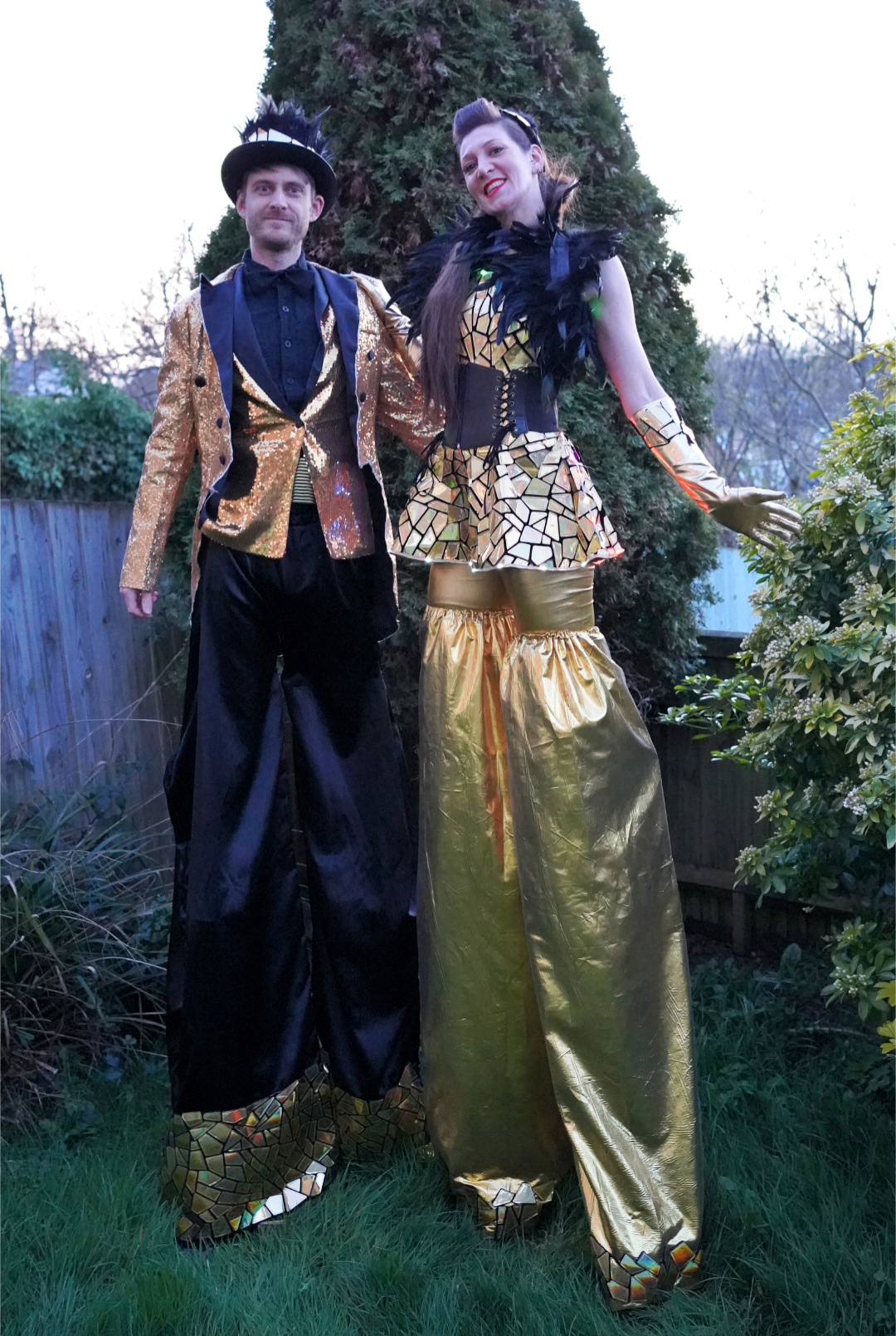 Two performers in gold mirrored costumes pose in a garden, with black feather accents, bright dusk light, and striking event-entertainment flair.