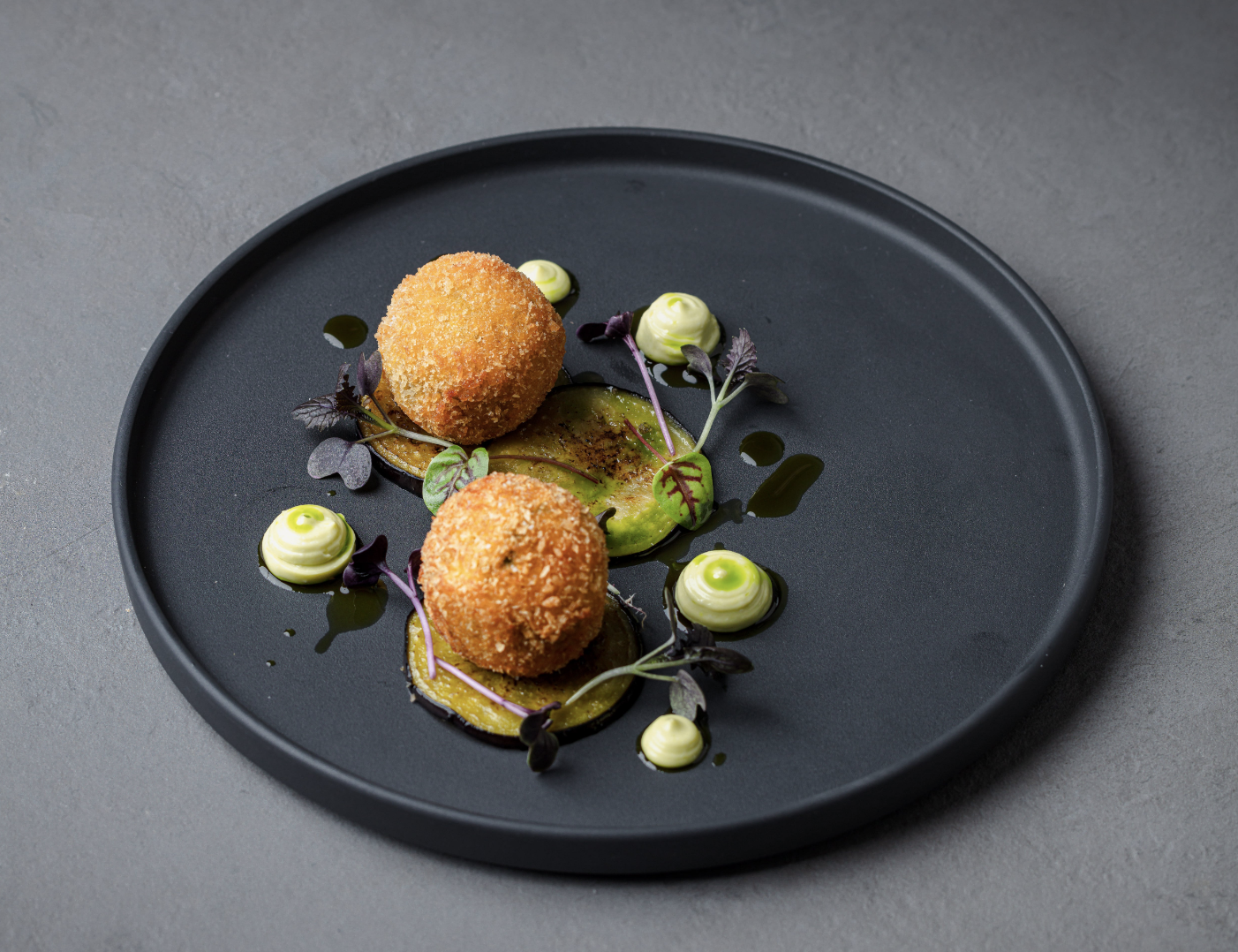 Golden, crisp croquettes are elegantly arranged on a matte black plate, garnished with vibrant green sauce and microgreens, exuding upscale dining vibes.