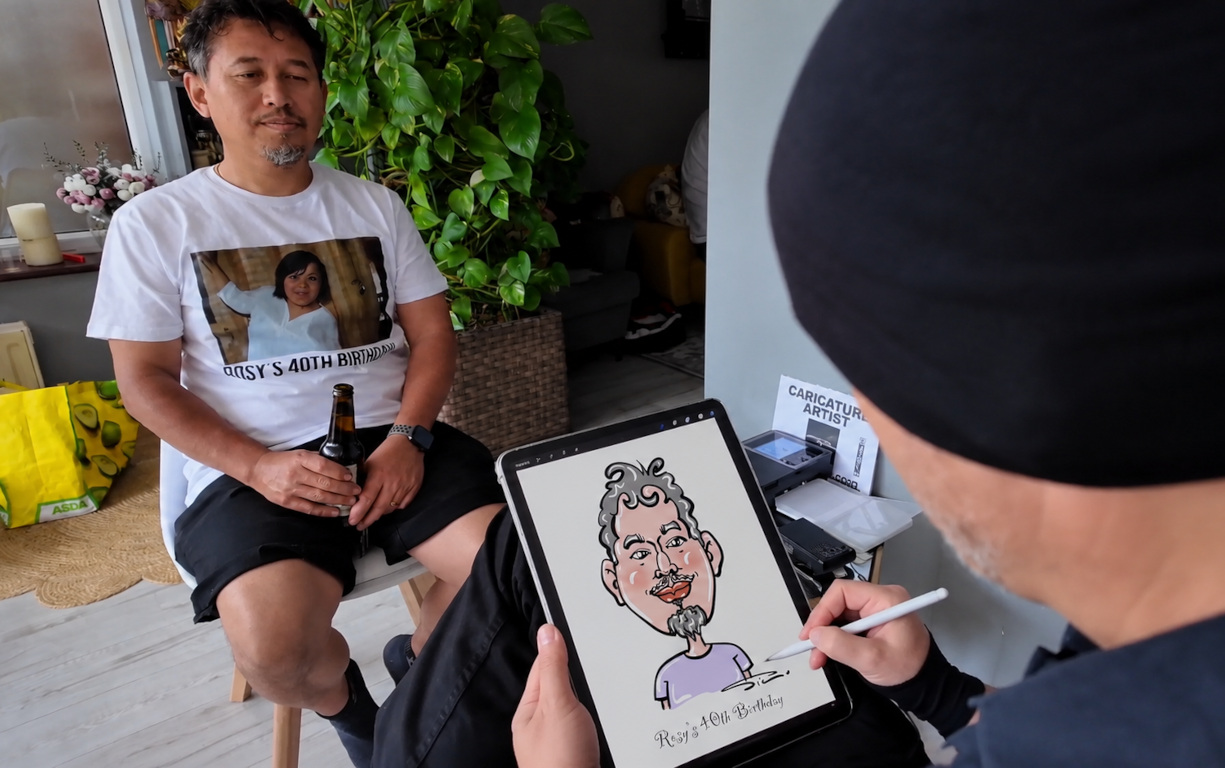 Live caricature artist at a 40th birthday, sketching on a tablet beside a seated guest; bright indoor light, casual catering vibe.
