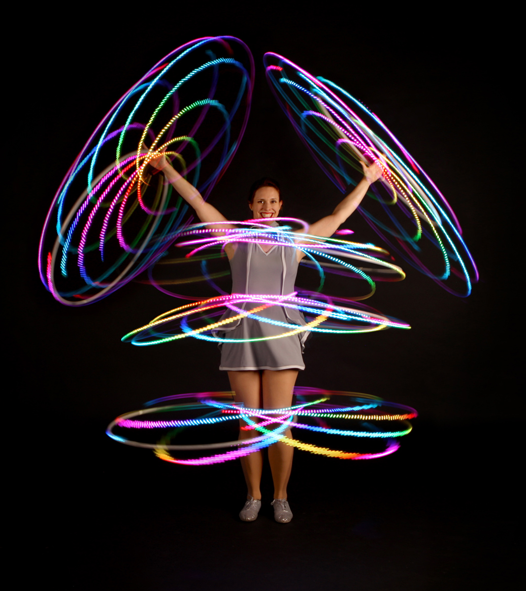 A lively performance showcases a smiling woman in a silver dress, energetically twirling colorful LED hoops against a dark backdrop.