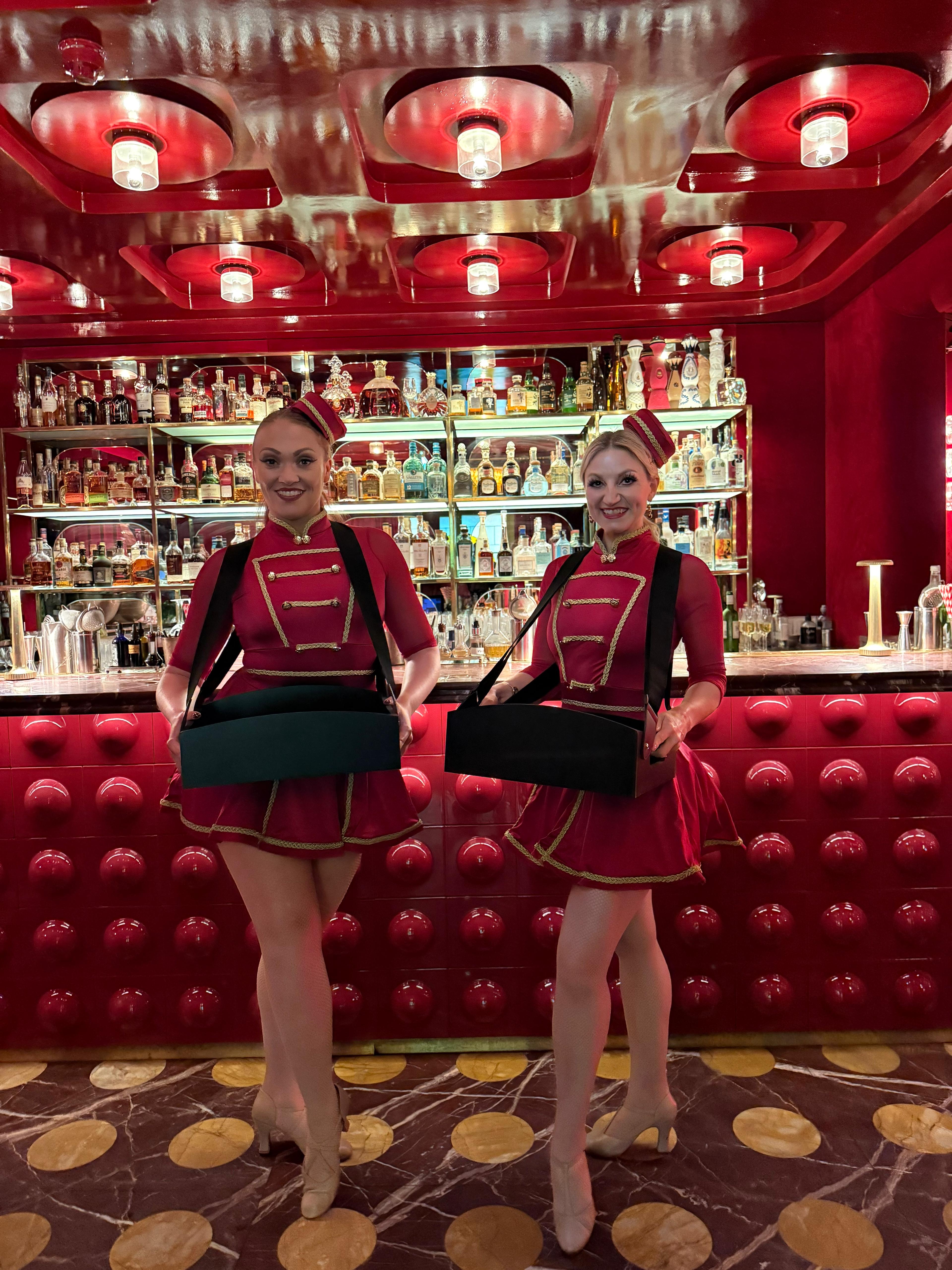 Two uniformed hosts stand before a red cocktail bar with backlit liquor shelves, polished brass, and dramatic ceiling lights.