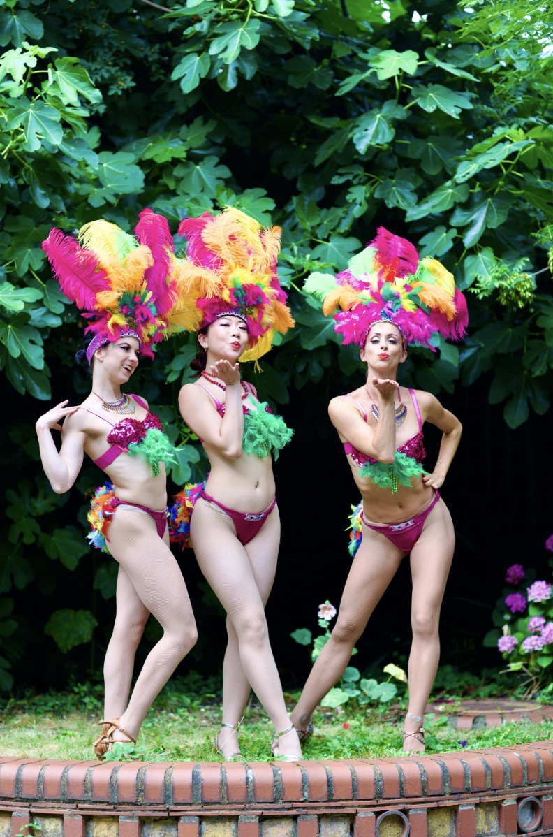 A vibrantly costumed performer with a feathered headdress poses playfully against a backdrop of lush greenery and brick, radiating festive energy.