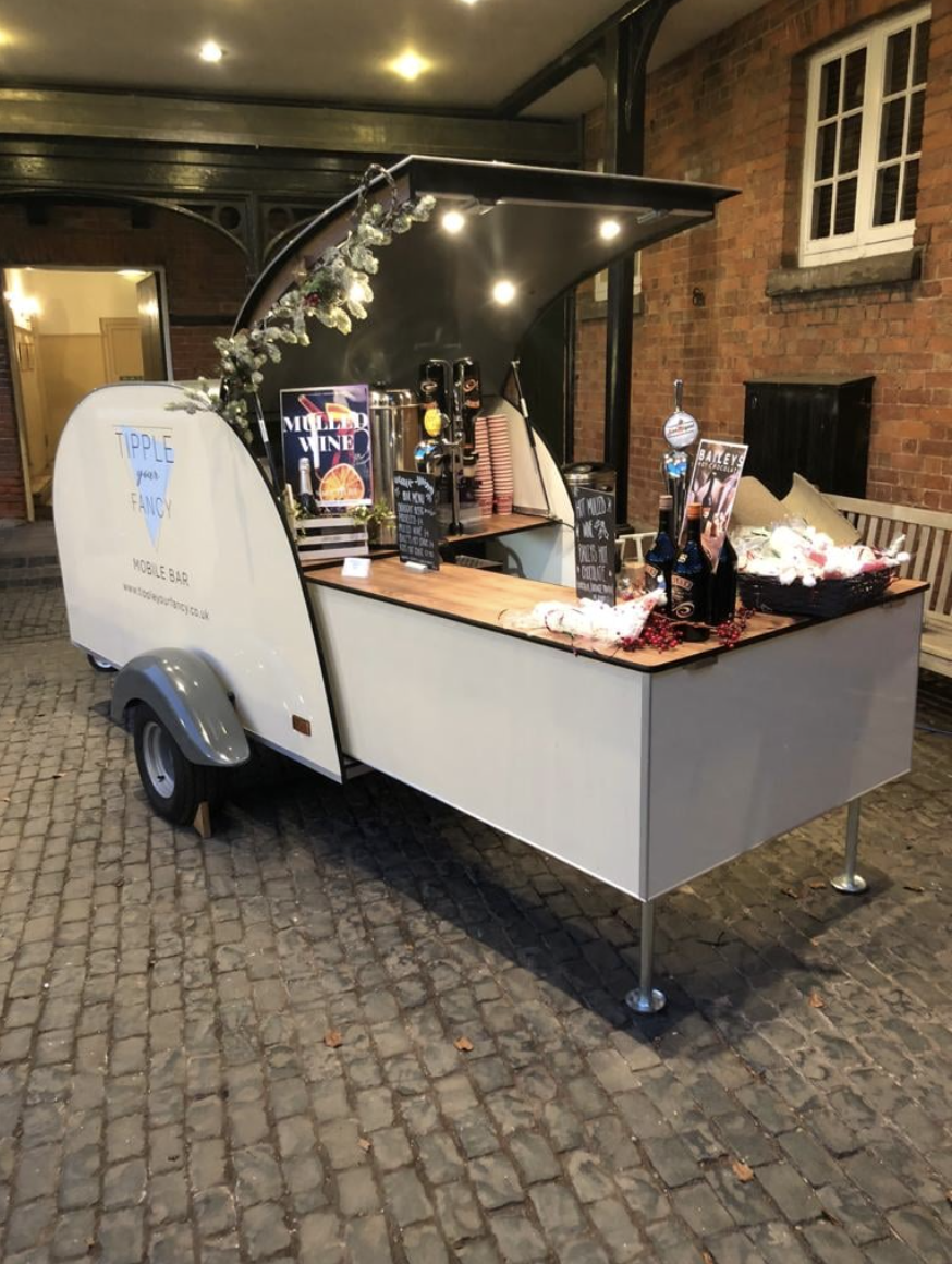 A stylish mobile bar featuring a wooden countertop, neatly arranged drinks, and festive decor, illuminated by soft ambient lighting for a cozy atmosphere.