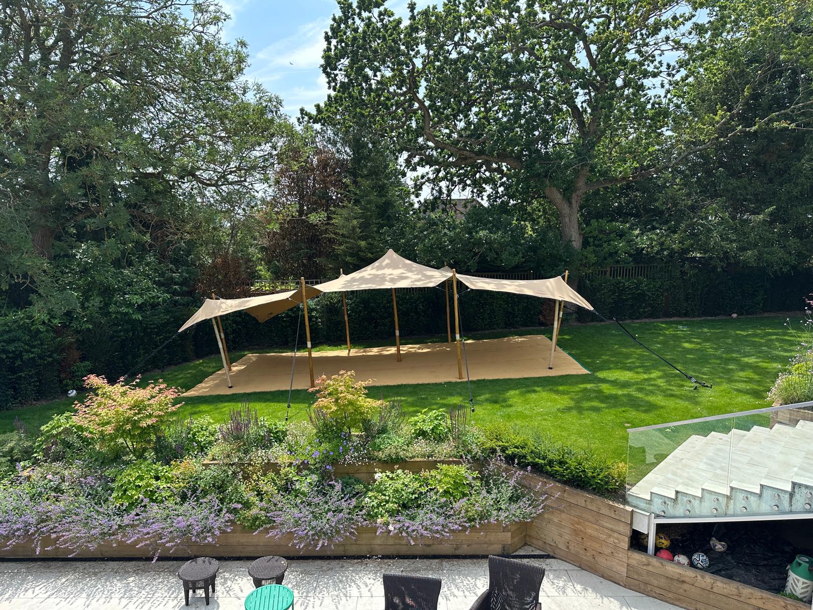 A spacious garden features a beige tent on a sandy area, surrounded by lush greenery and vibrant flowers, under bright daylight.