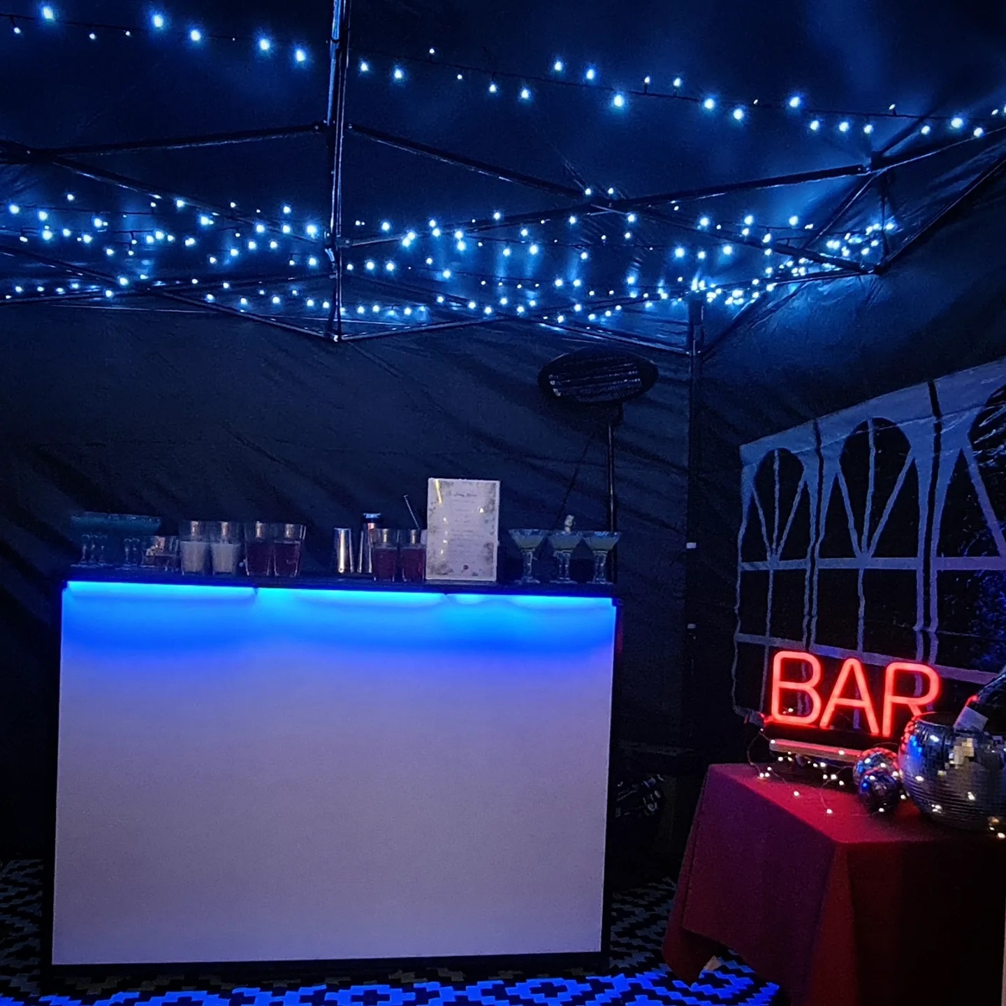 A sleek bar with a glowing blue LED base showcases clear glasses filled with colorful drinks, under twinkling fairy lights for a festive vibe.