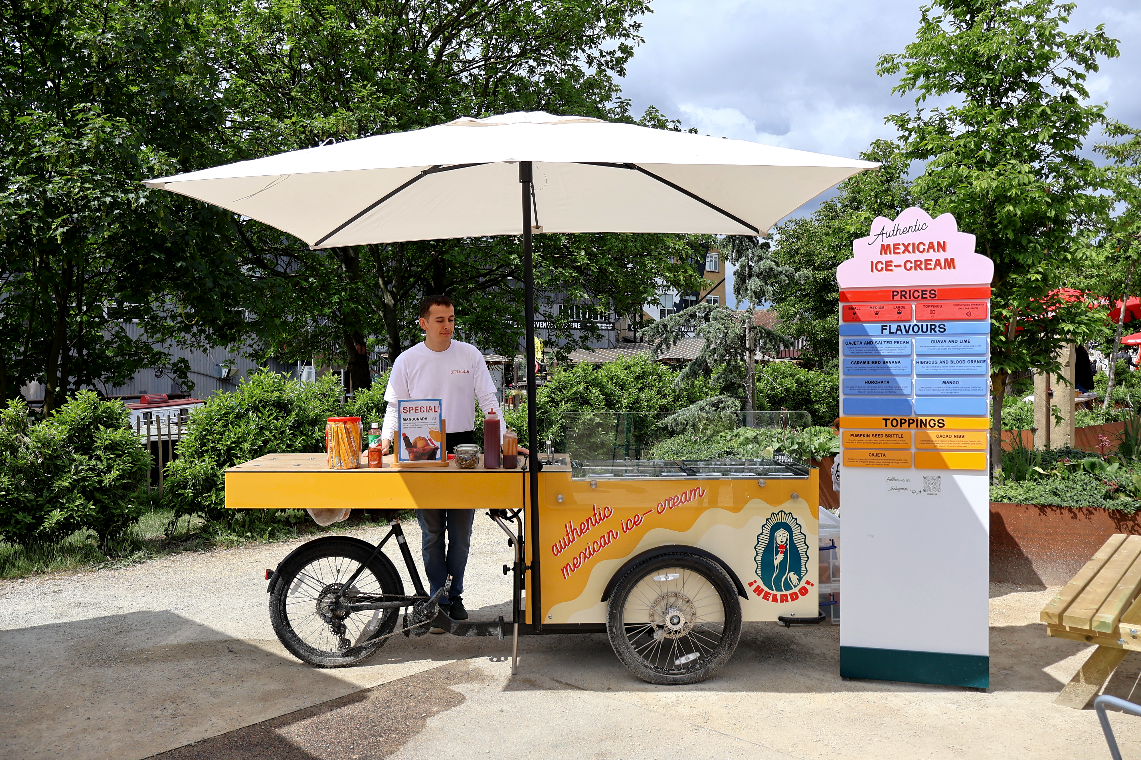 A vibrant yellow ice-cream cart with glass display holds various toppings, surrounded by lush greenery, radiating a cheerful daytime vibe.