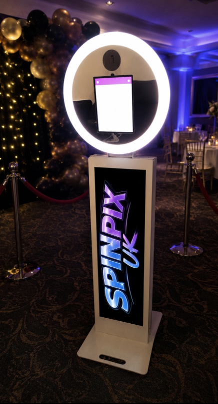 A glowing SpinPix UK photo booth stands on a patterned carpet beside black-gold balloon decor, under moody purple lighting at a stylish catering event.