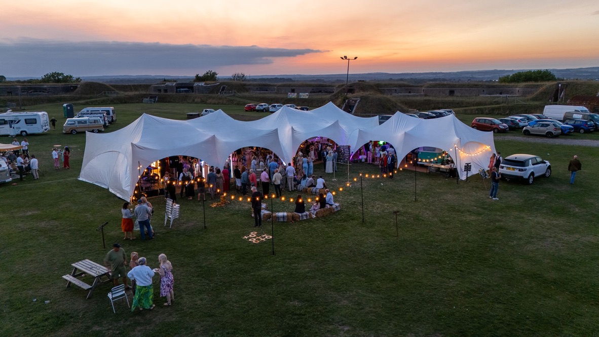 A lively outdoor catering event under a large white tent, illuminated by string lights at sunset, featuring guests mingling on green grass.