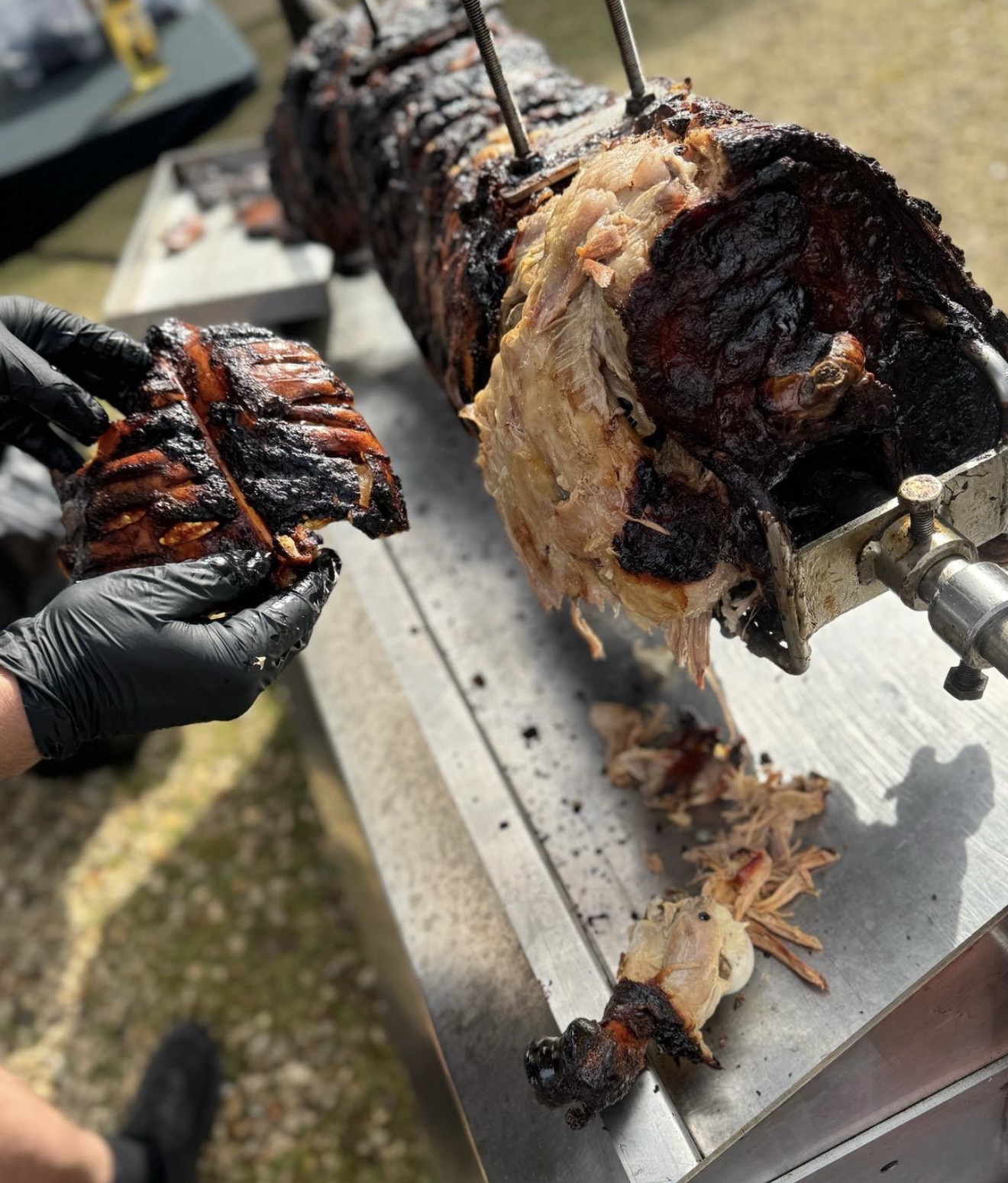 Gloved caterers carve crispy rotisserie pork over a stainless buffet station, with charred crackling, warm daylight, and polished outdoor service.