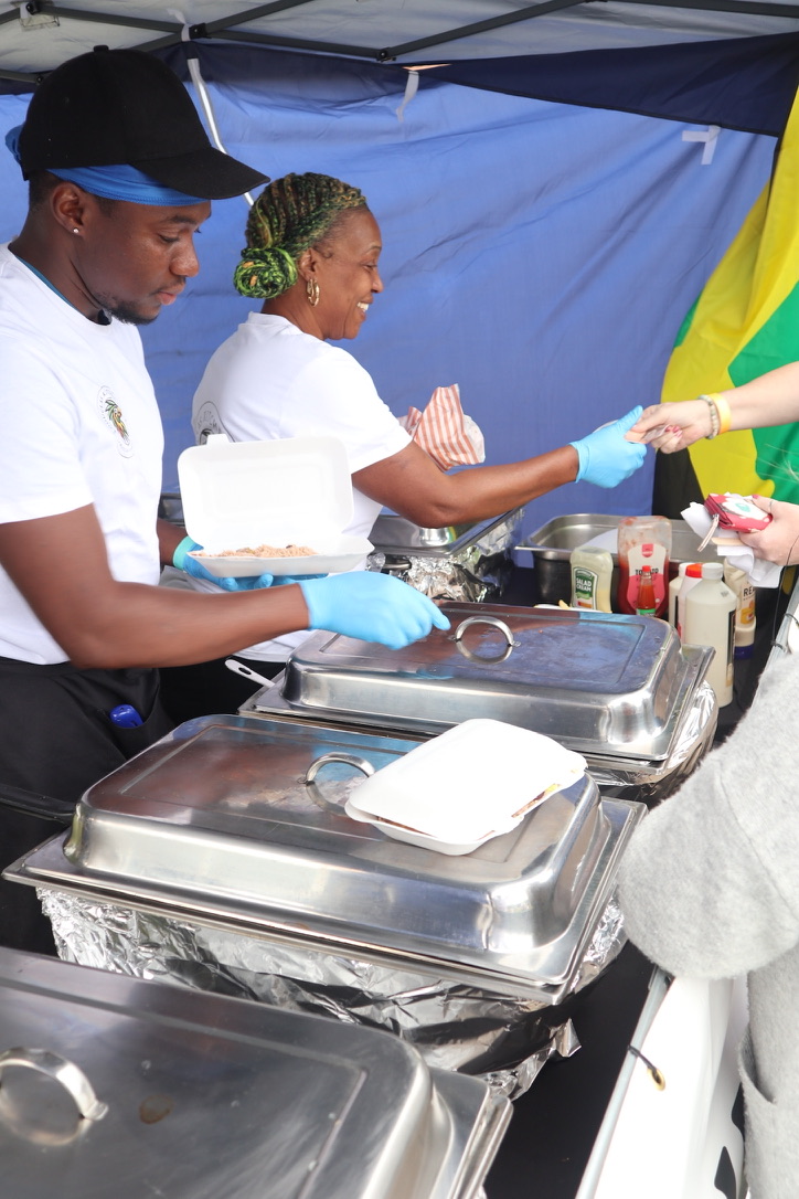 Chefs serve a catering buffet under a blue canopy, with polished stainless chafing dishes, sauces and gloves in bright daylight.