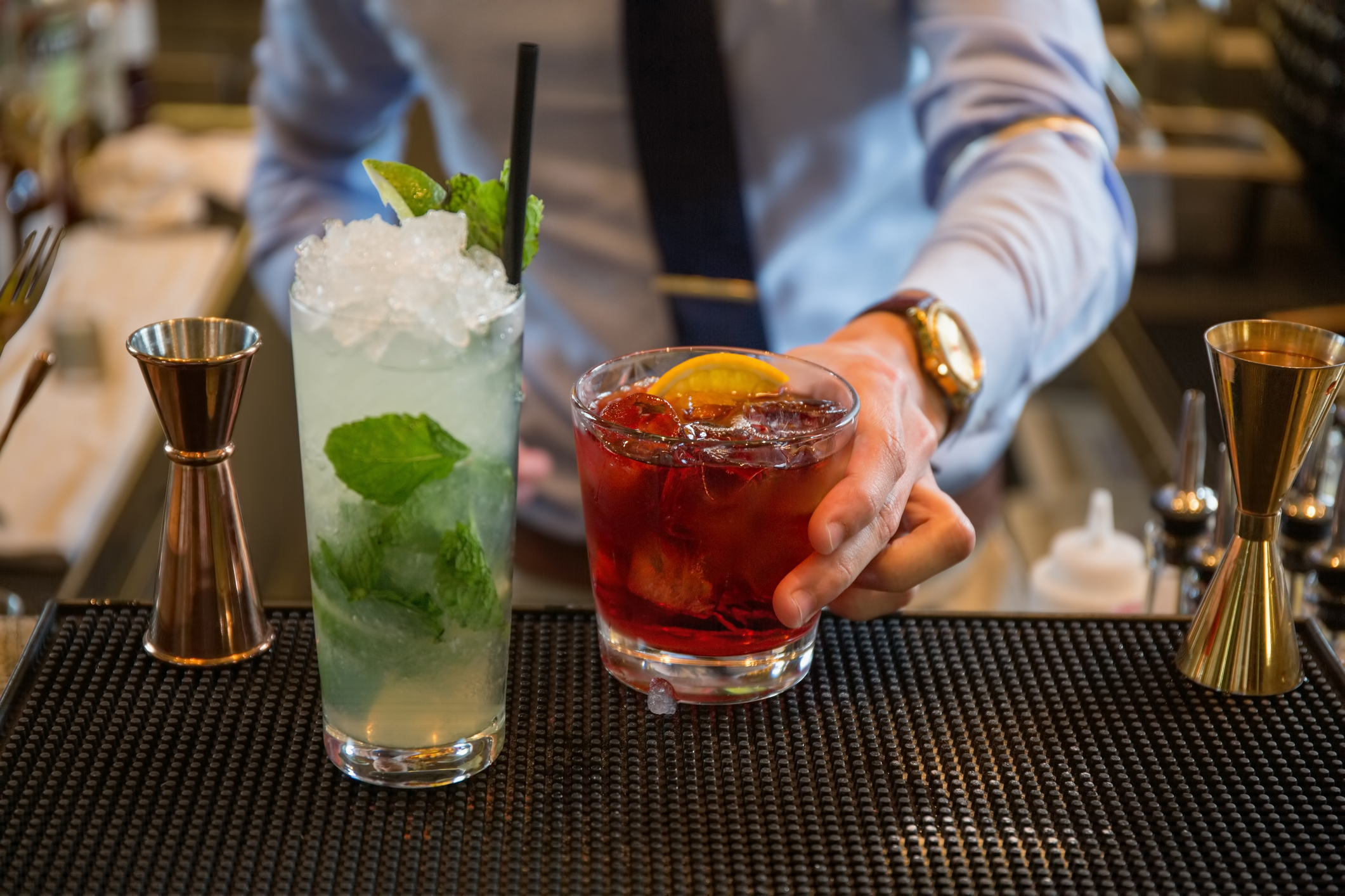 A bartender presents a refreshing minty mojito and a vibrant bourbon cocktail, highlighting expert mixology amidst a sophisticated bar setup.