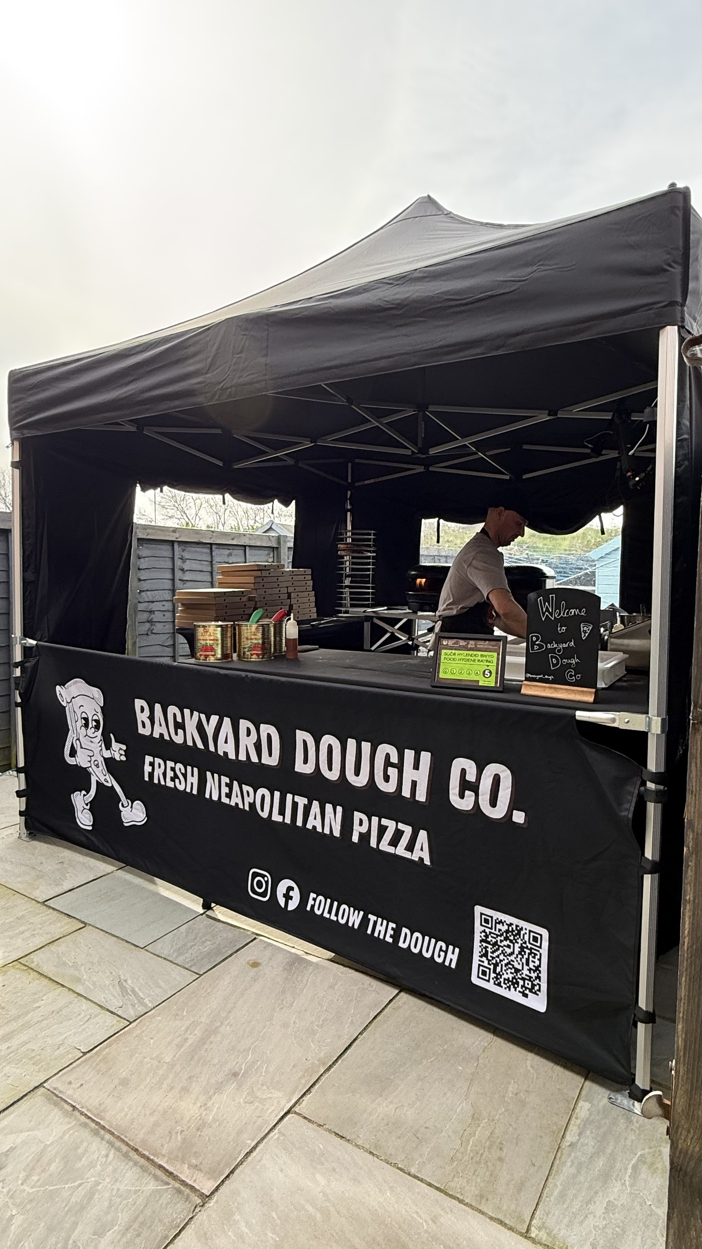 A vibrant outdoor pizza stall features a black canopied setup, showcasing stacks of pizza boxes and colorful condiments amidst warm daylight.