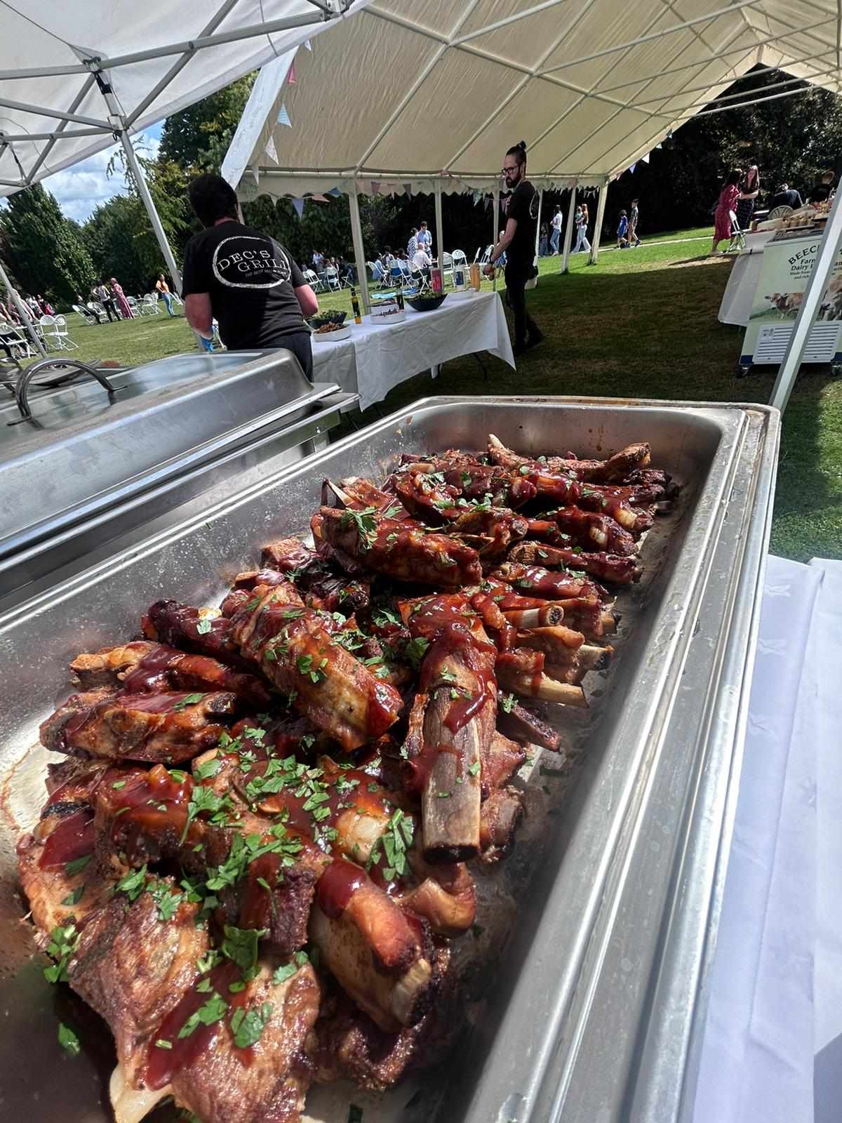Buffet-style catering under a bright marquee, with polished chafing trays of glazed ribs, parsley garnish, white linens, and a relaxed outdoor crowd.
