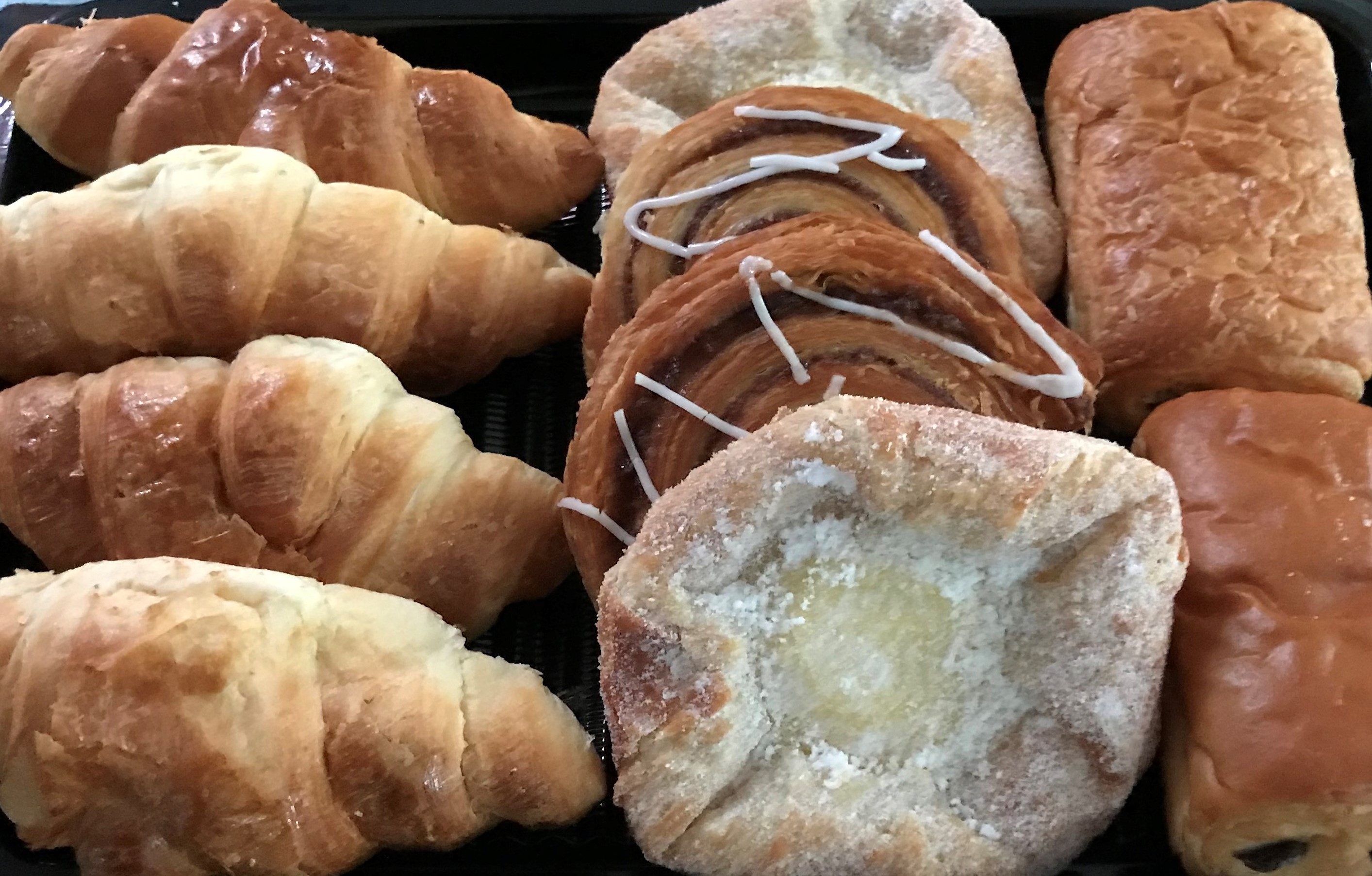 Golden croissants, sweet rolls, and danishes arranged neatly on a black tray under soft lighting, showcasing artful pastry craftsmanship.