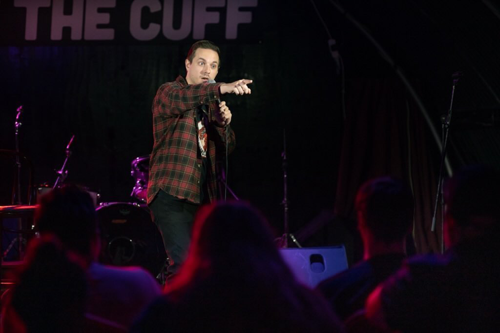Comedian in red plaid shirt points on a dark club stage, with purple-red lighting, microphone, drums, and a small crowd creating energetic live entertainment vibes.