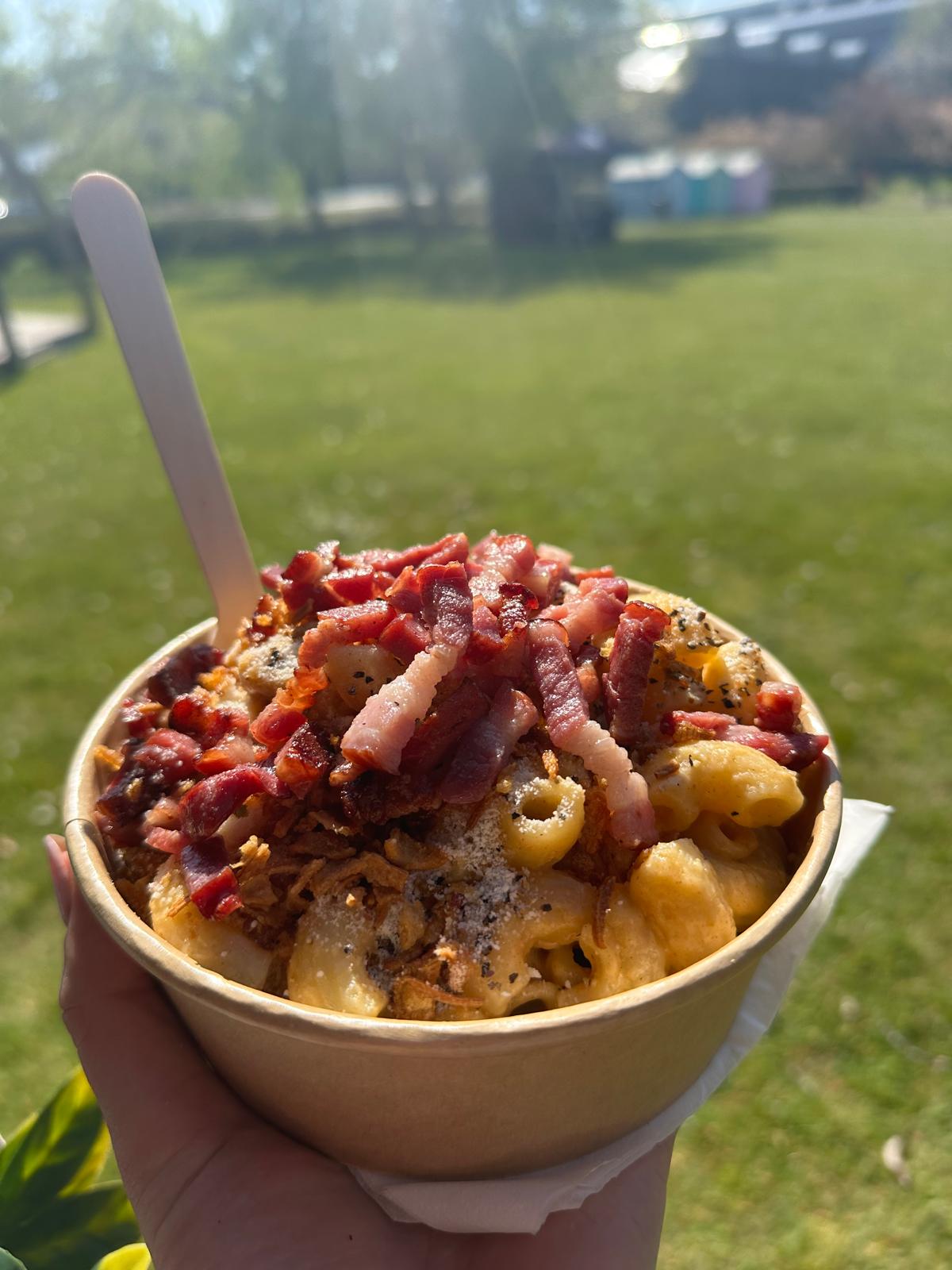 A vibrant bowl of creamy macaroni topped with crispy bacon, pepper, and crunchy onion flakes, set against sunny outdoor greenery.