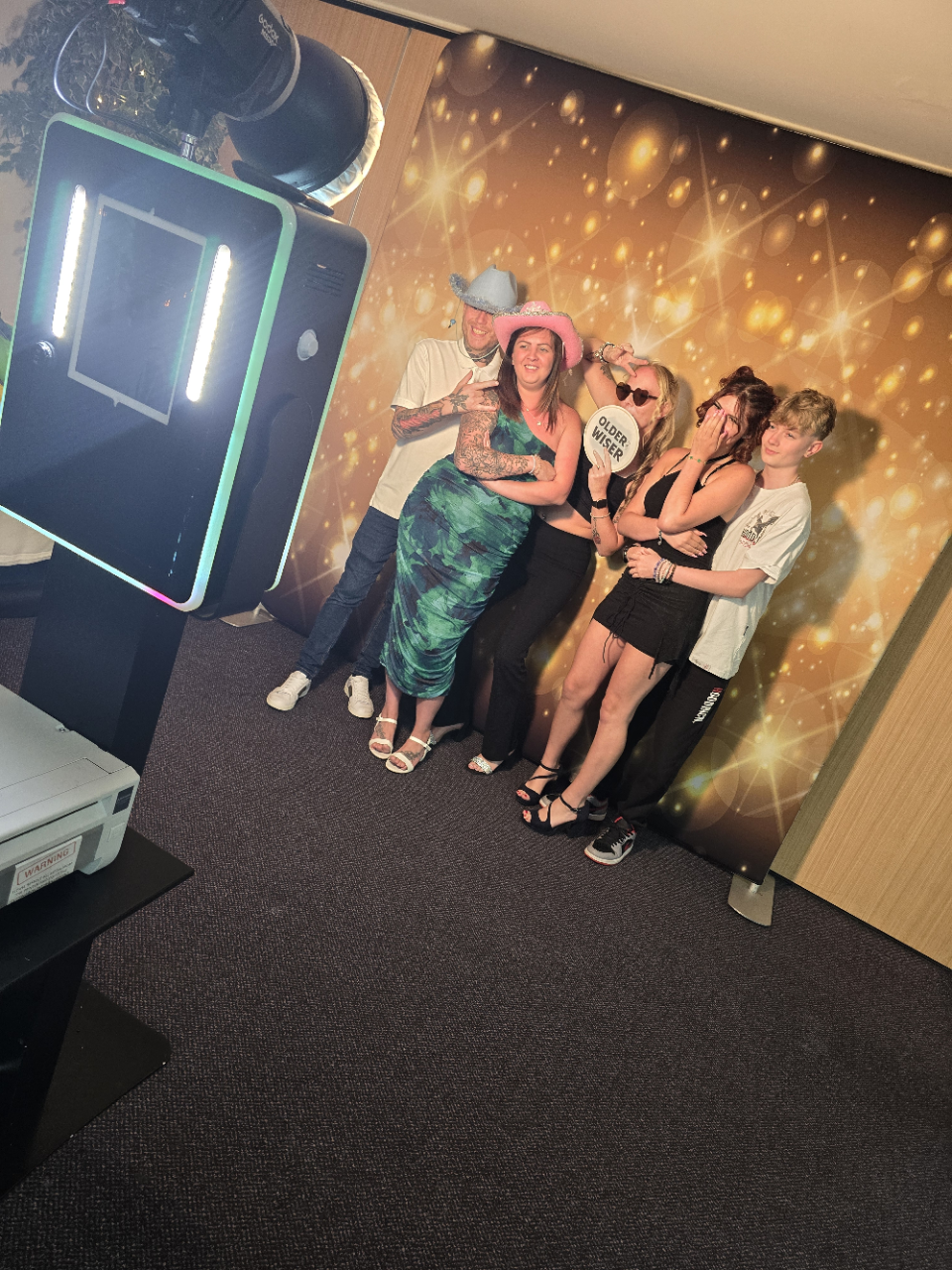 A lively group poses playfully in front of a sparkling gold backdrop with studio lighting, showcasing hats and cheerful expressions.