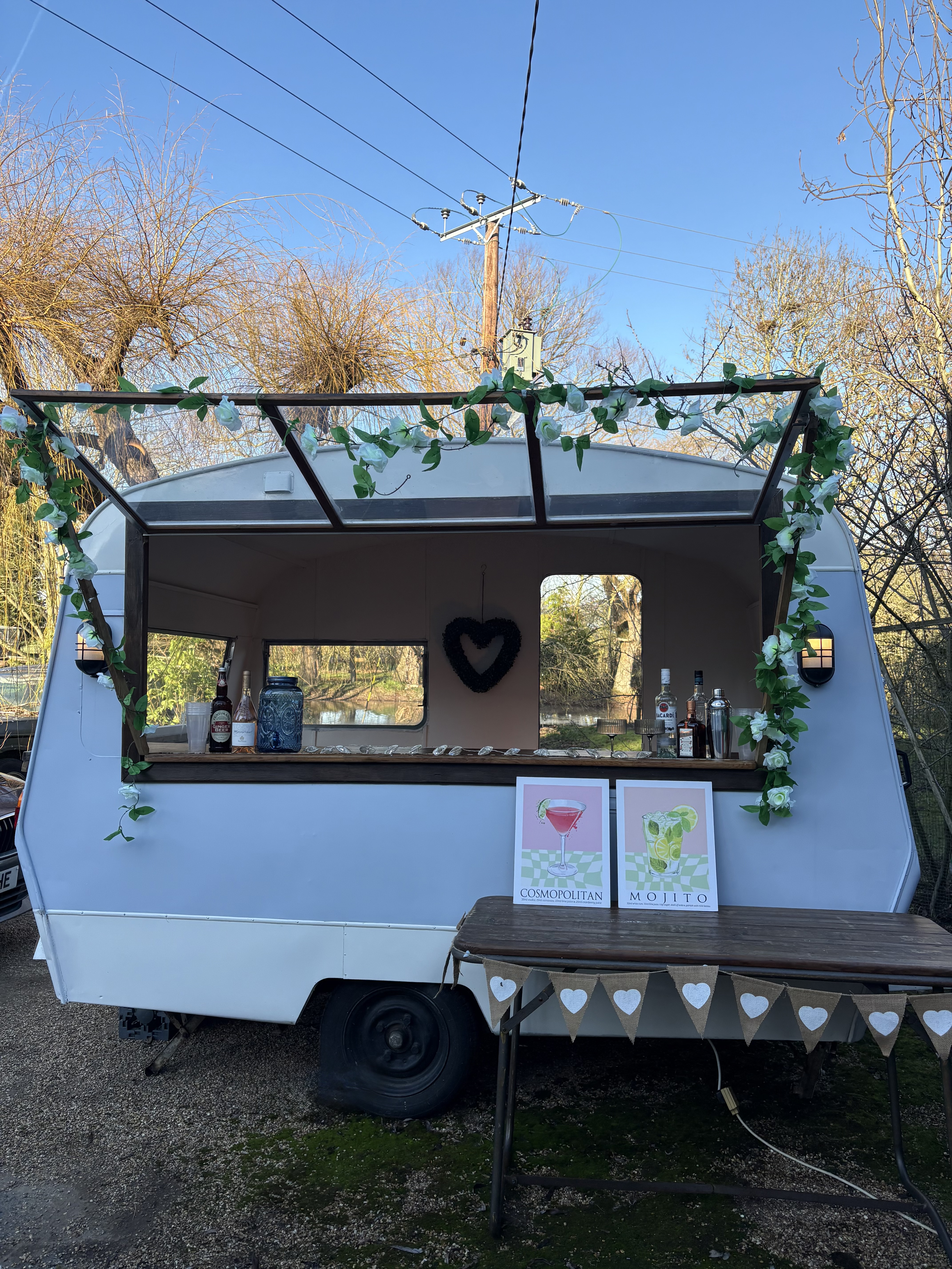 A vintage mobile bar displays cocktail choices, with lush greenery adorning its frame. Bright daylight enhances the inviting atmosphere.
