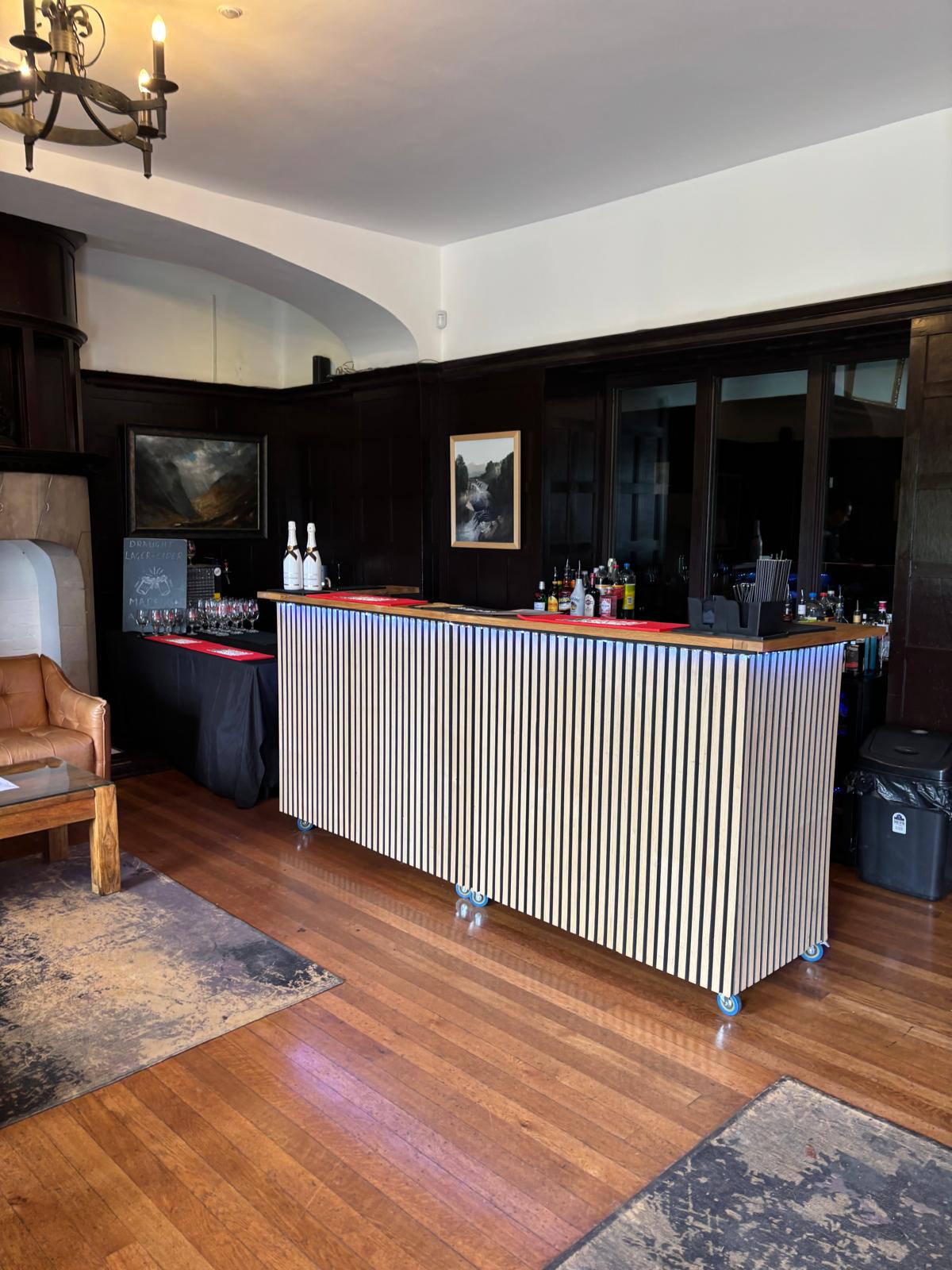 Sophisticated catering bar with striped wood front, glassware, bottles, and red mats, set on polished hardwood under warm chandelier light.