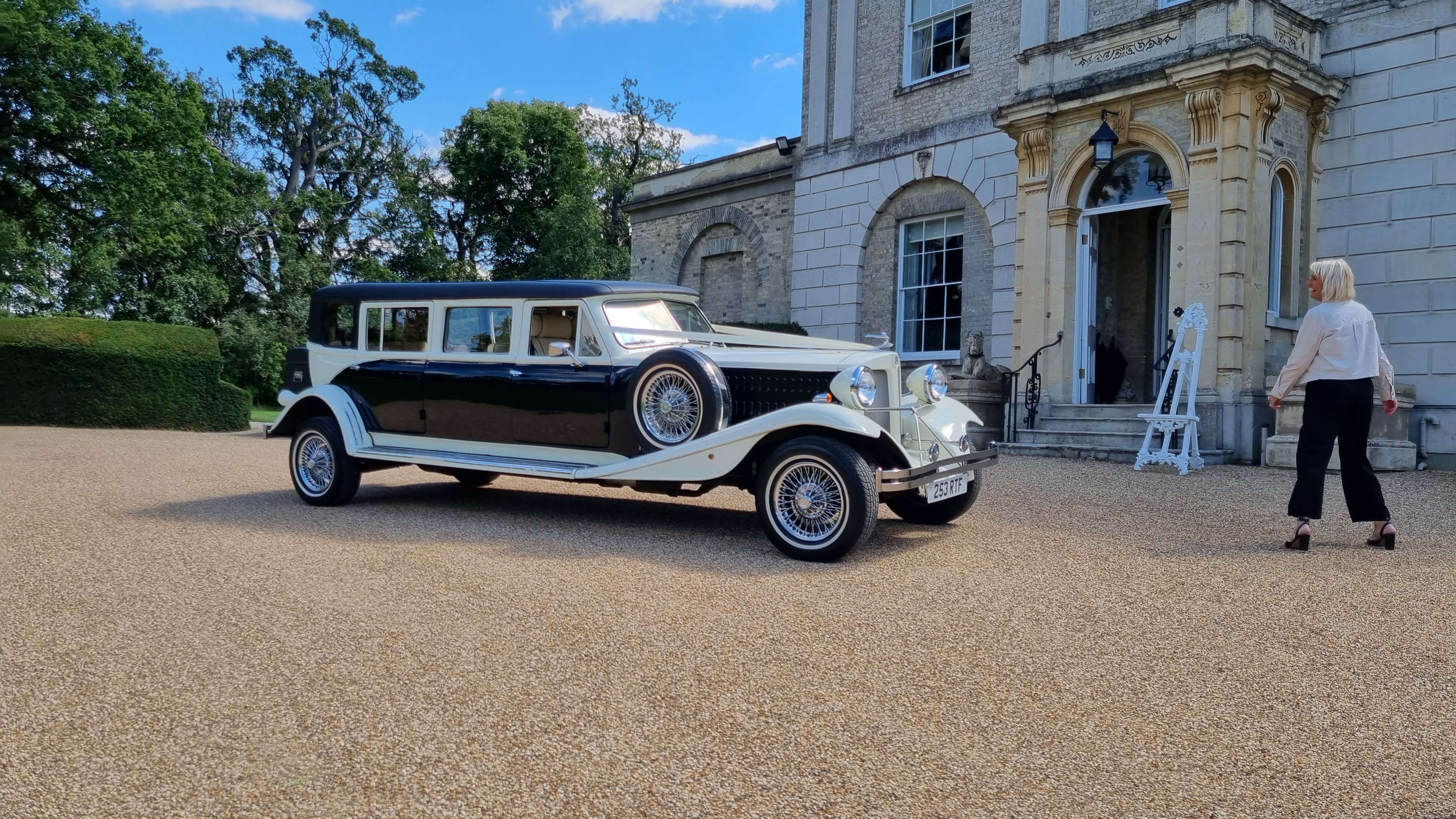 A vintage black and cream limousine parked on gravel, in front of a grand estate with lush greenery, providing an elegant atmosphere.