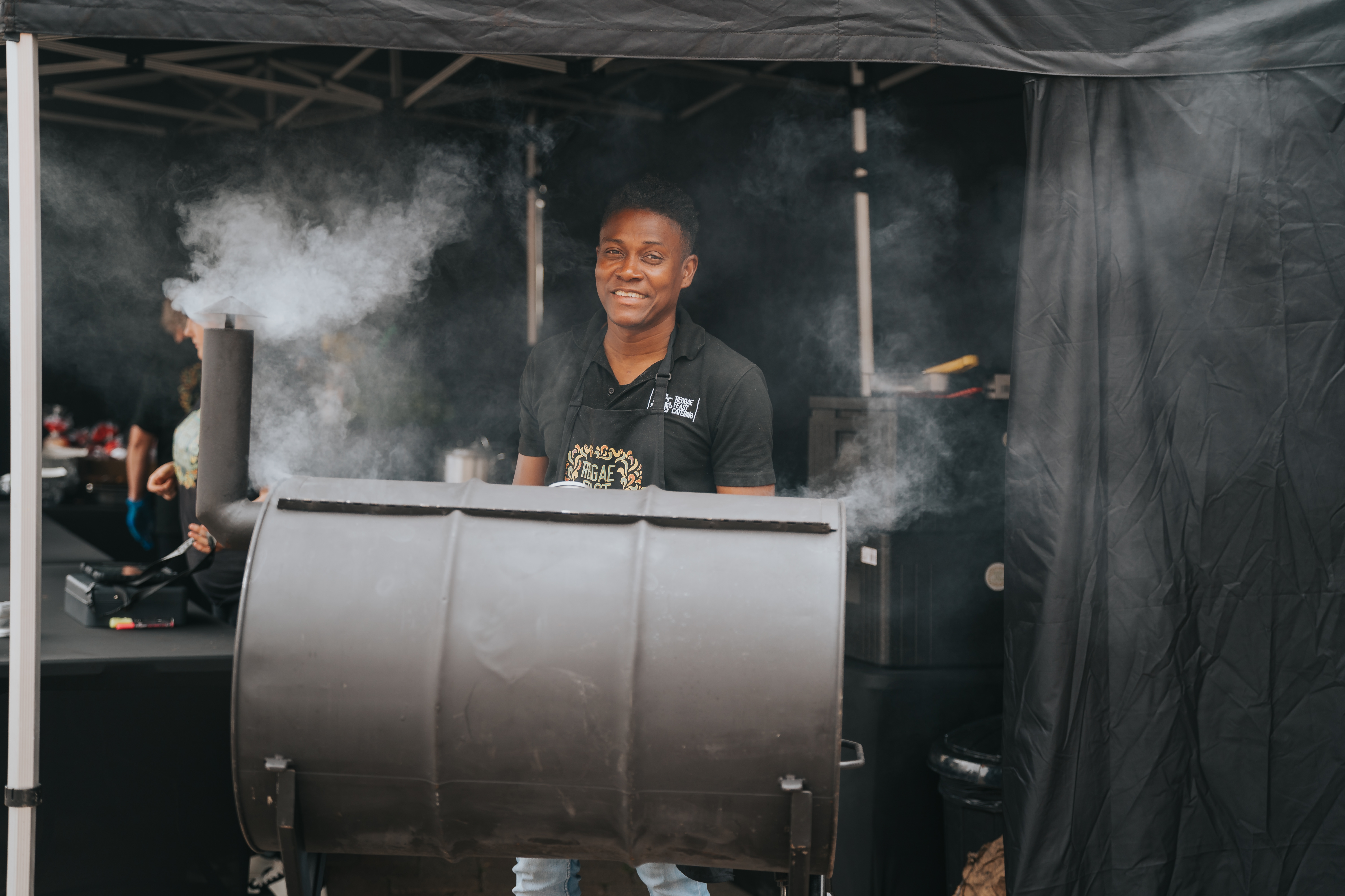 Smiling caterer at a smoky black barbecue smoker under a tent, with polished setup and warm outdoor event-service vibes.