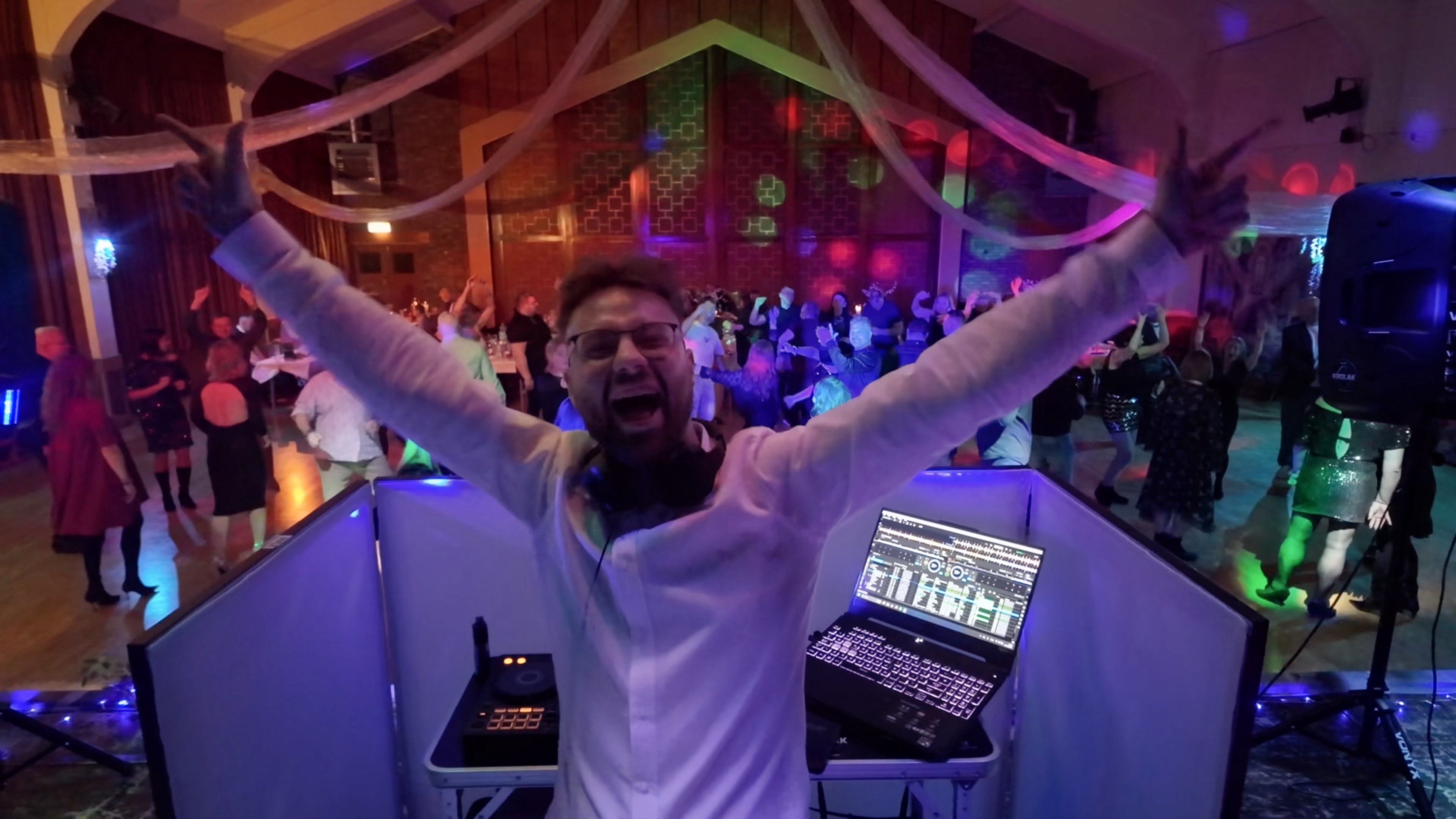 Energetic wedding DJ booth with laptop, controller and speaker, neon lighting, cheering crowd, draped hall, polished event catering vibe.