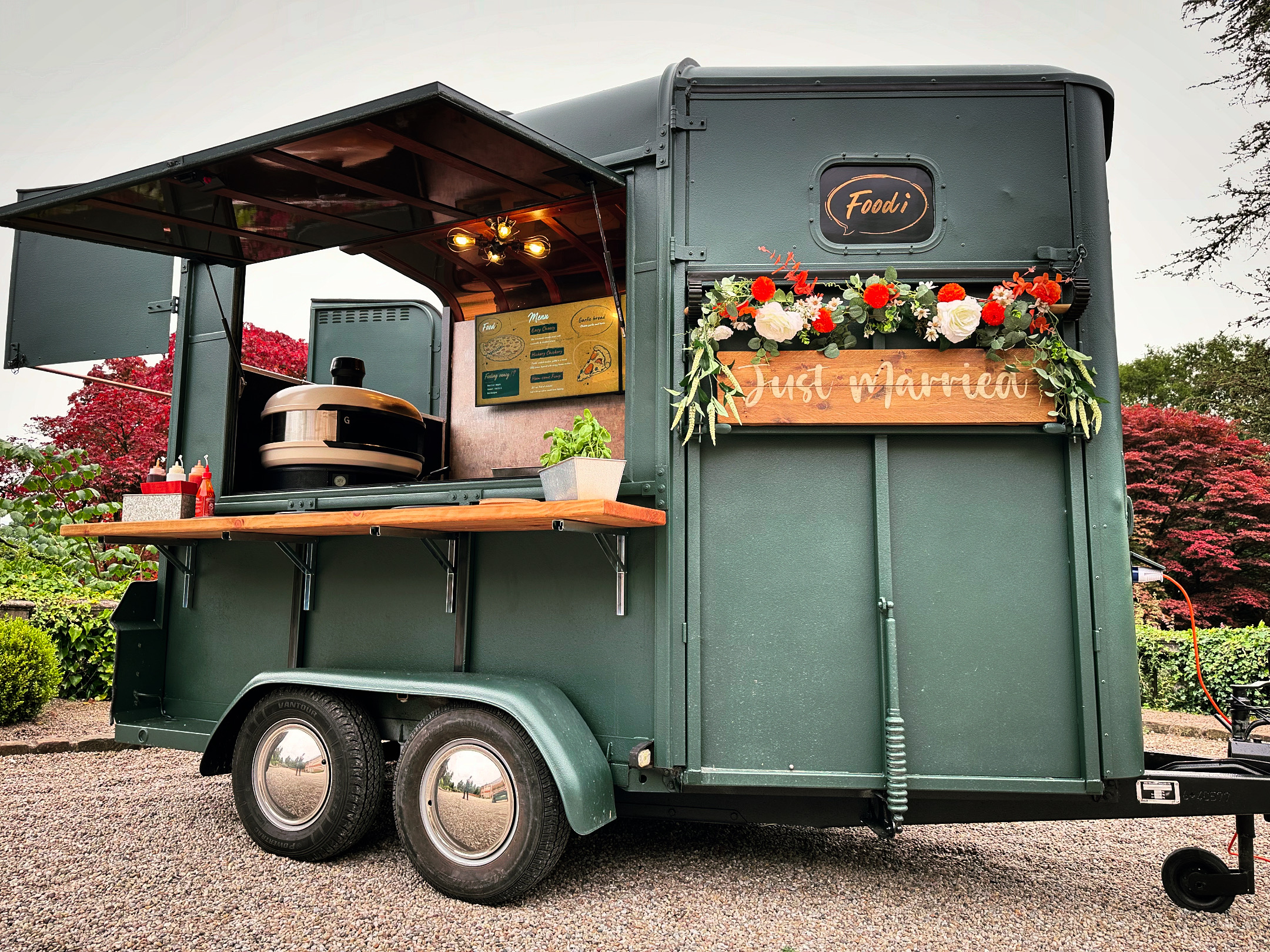 A vintage green food trailer features a polished wooden countertop, vibrant floral decor, and a welcoming menu display, set in a serene garden.